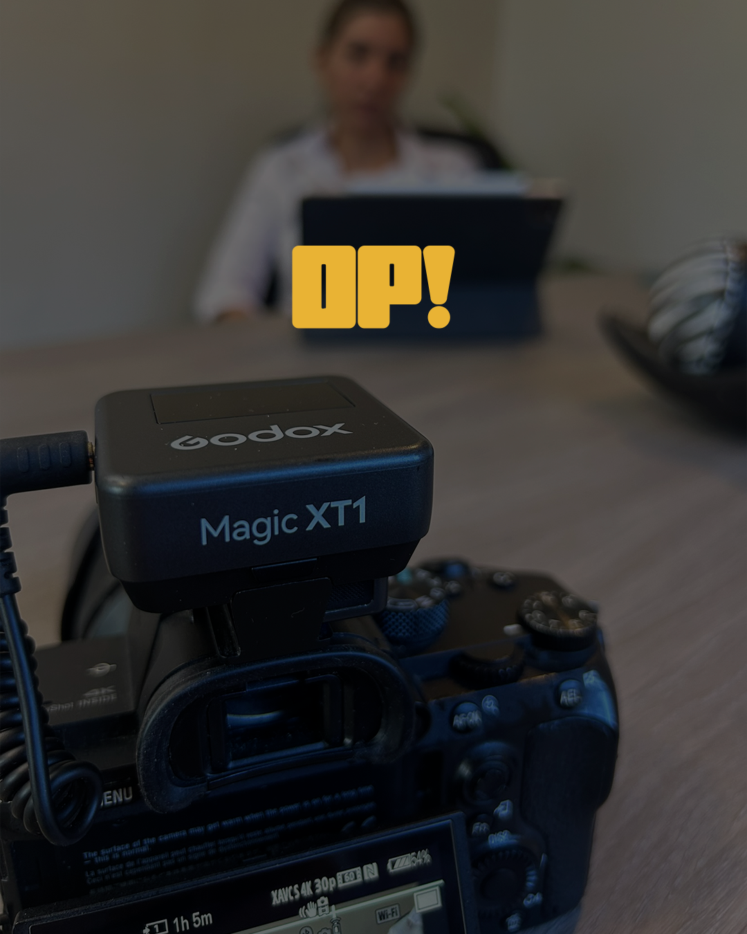 Una cámara profesional en primer plano con el objetivo enfocado en la pantalla, en un entorno de oficina con una mujer borrosa en el fondo, sentada en una mesa y sosteniendo una tablet, con la palabra 'UP!' en texto grande y amarilla en el centro de la imagen.