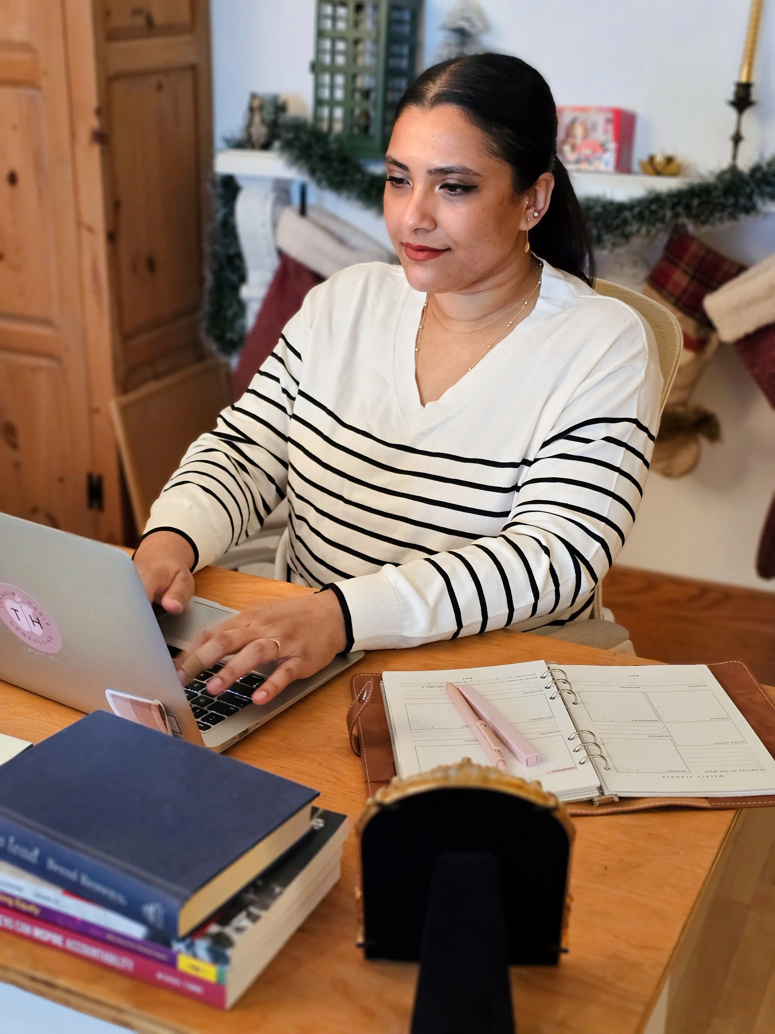 Woman working on a laptop at a wooden desk surrounded by books, a planner with a pink pen, and a stand in a cozy, decorated room.