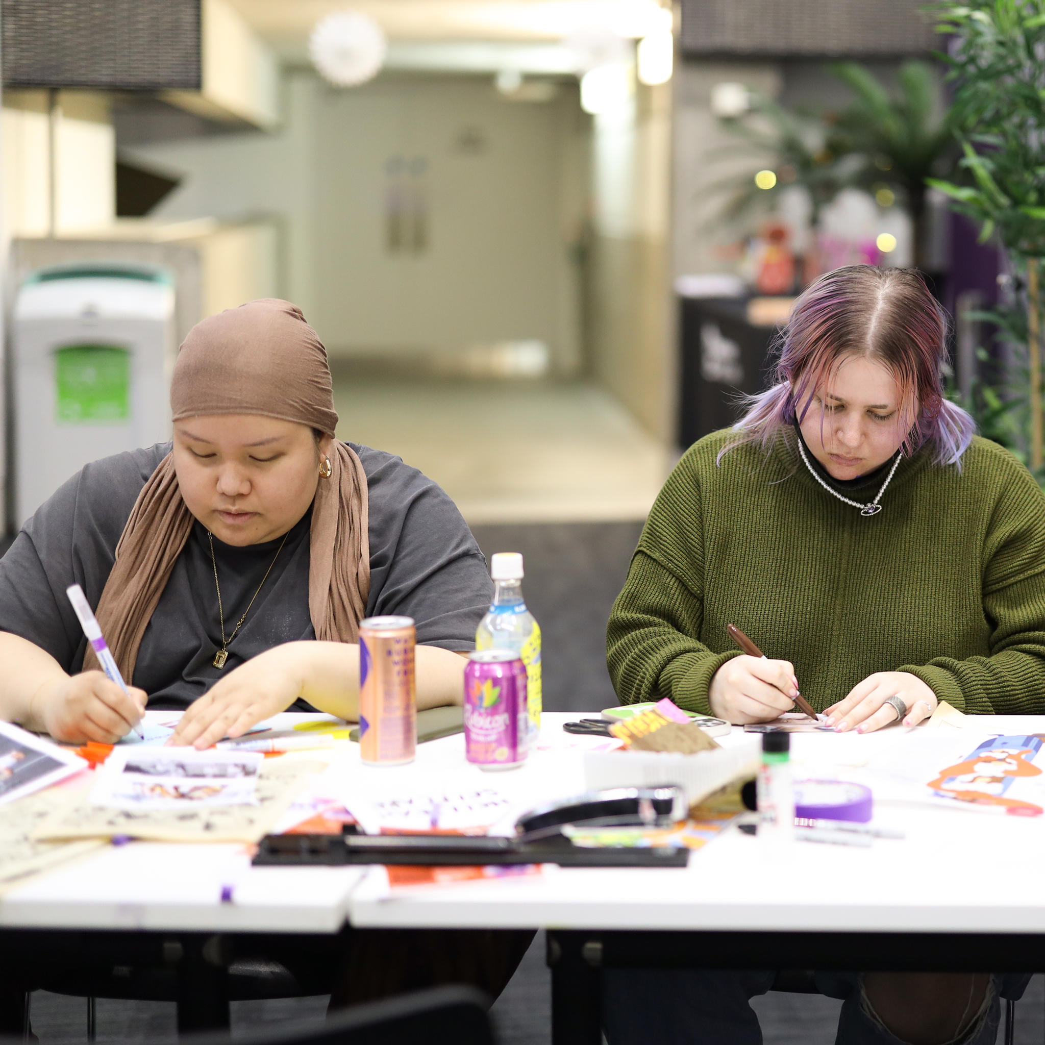 Two women sitting at a table working on craft or art projects with various supplies, drinks, and papers in front of them.