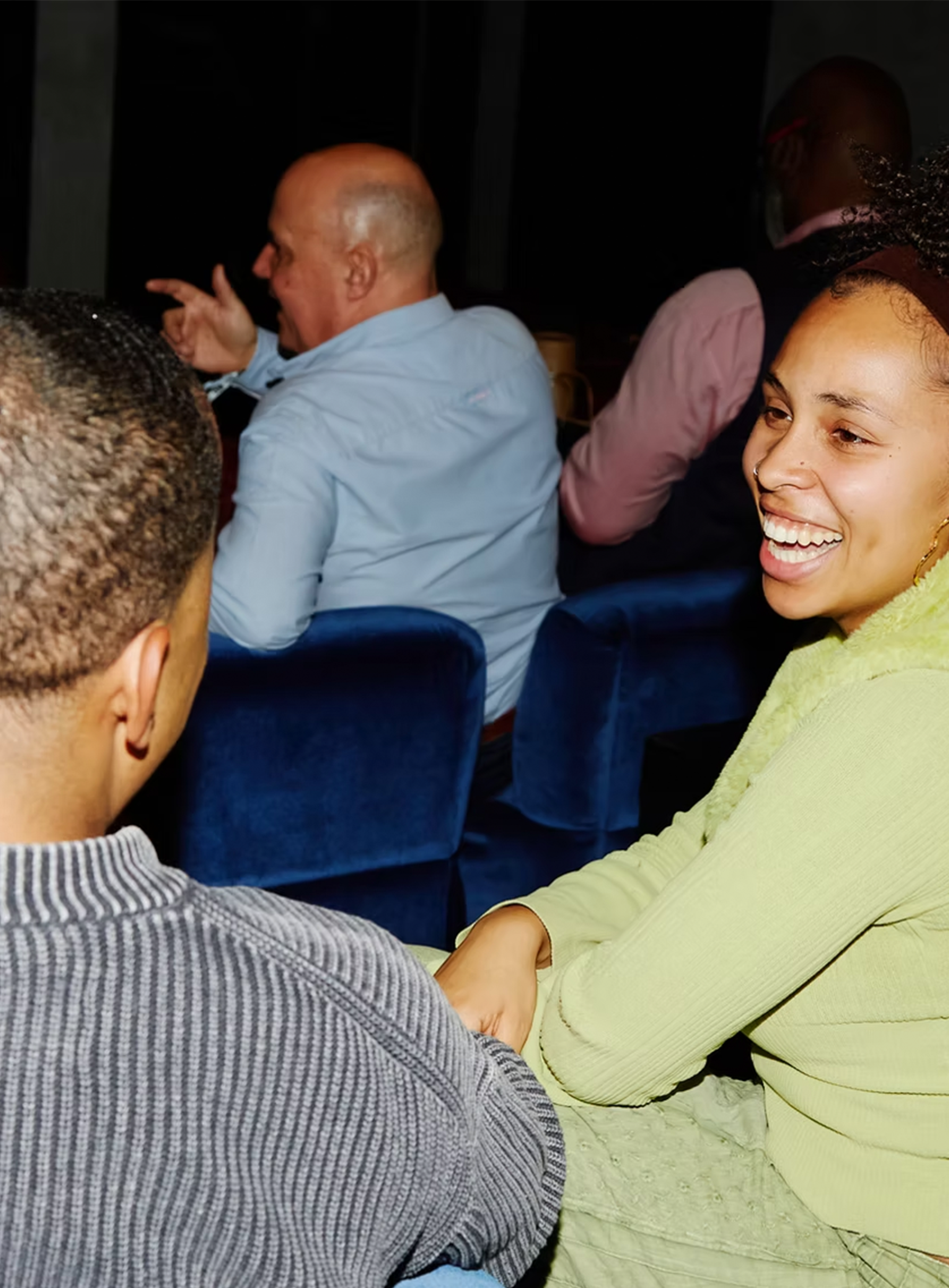 A woman in a yellow sweater smiling and holding hands with a man in a striped sweater during a conversation, with others seated behind them in a dimly lit setting.