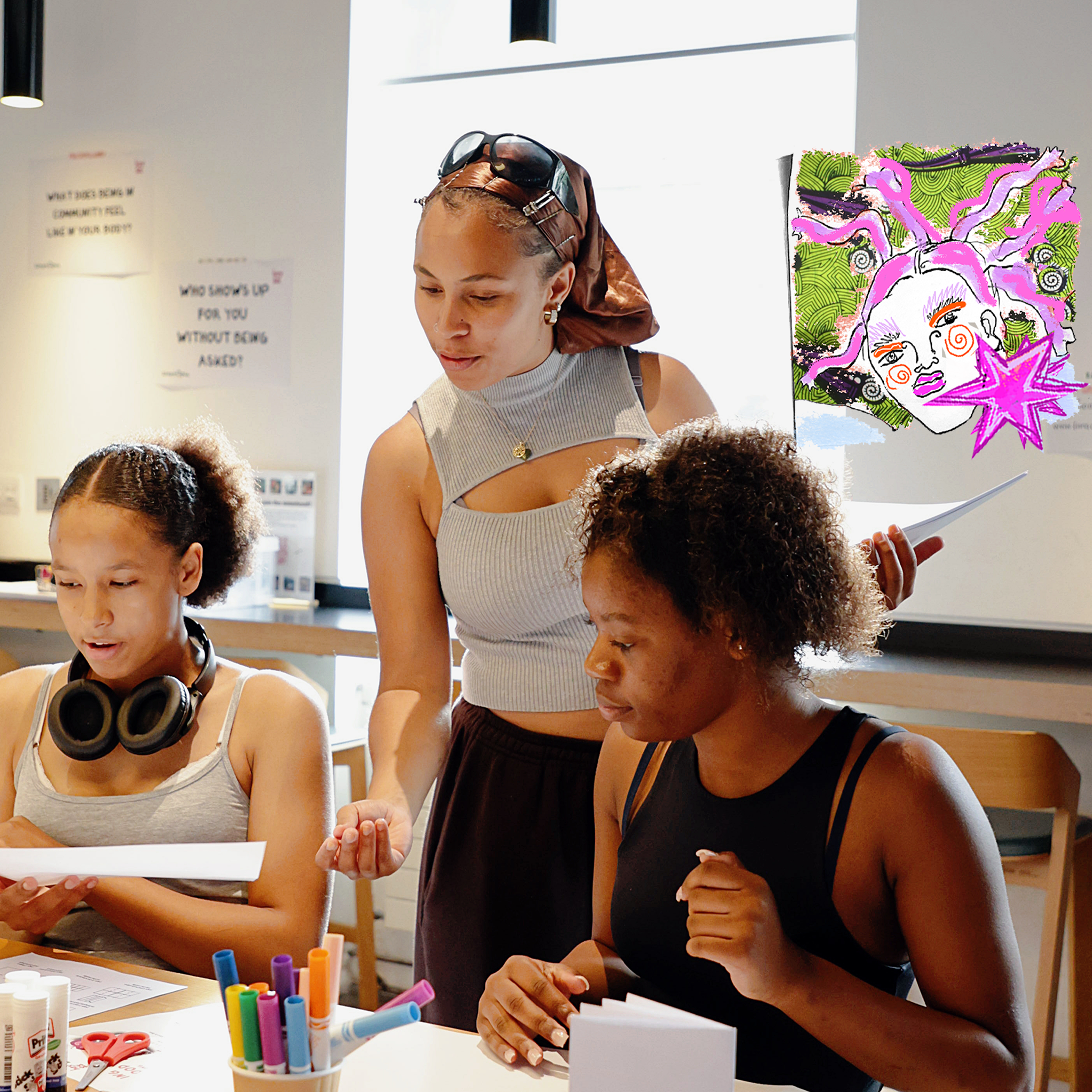 Three women in a classroom setting. Two are seated at a table, looking at papers, while the standing woman leans over them, engaging in conversation. Art supplies are visible on the table, and a colorful drawing of a woman's face with pink and purple hair is on the wall in the background.
