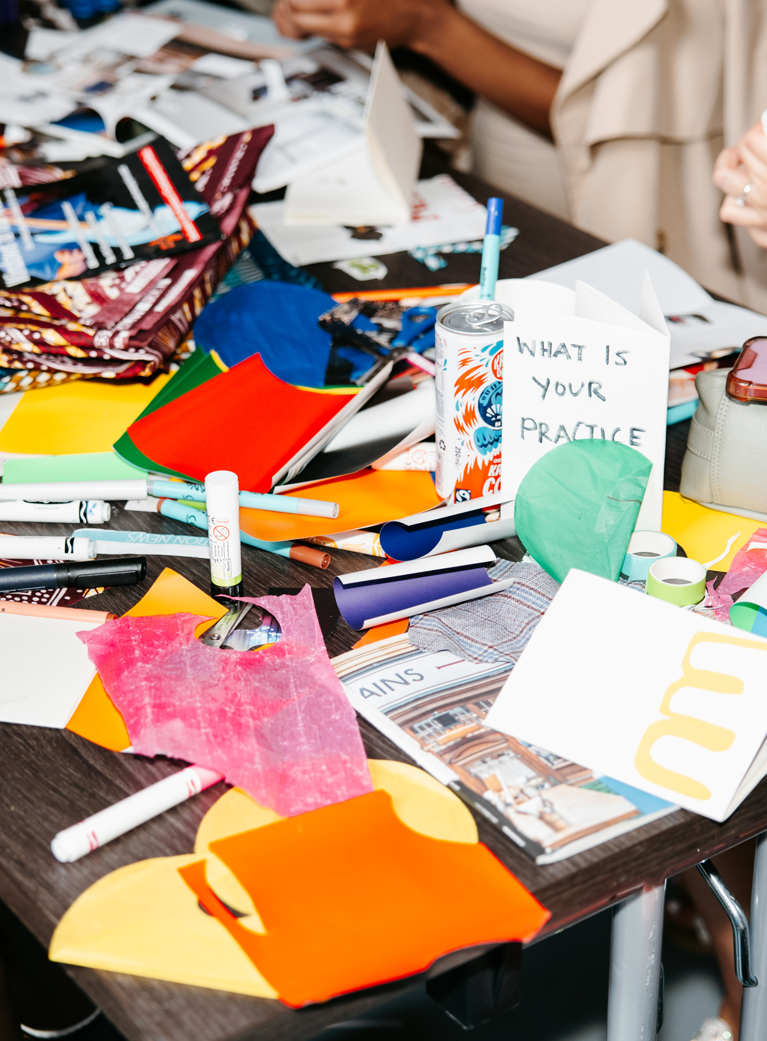 A cluttered table with various arts and crafts supplies, including paper, markers, rolls of tape, and a can of soda, with a sign reading 'What is your practice?' and people working in the background.