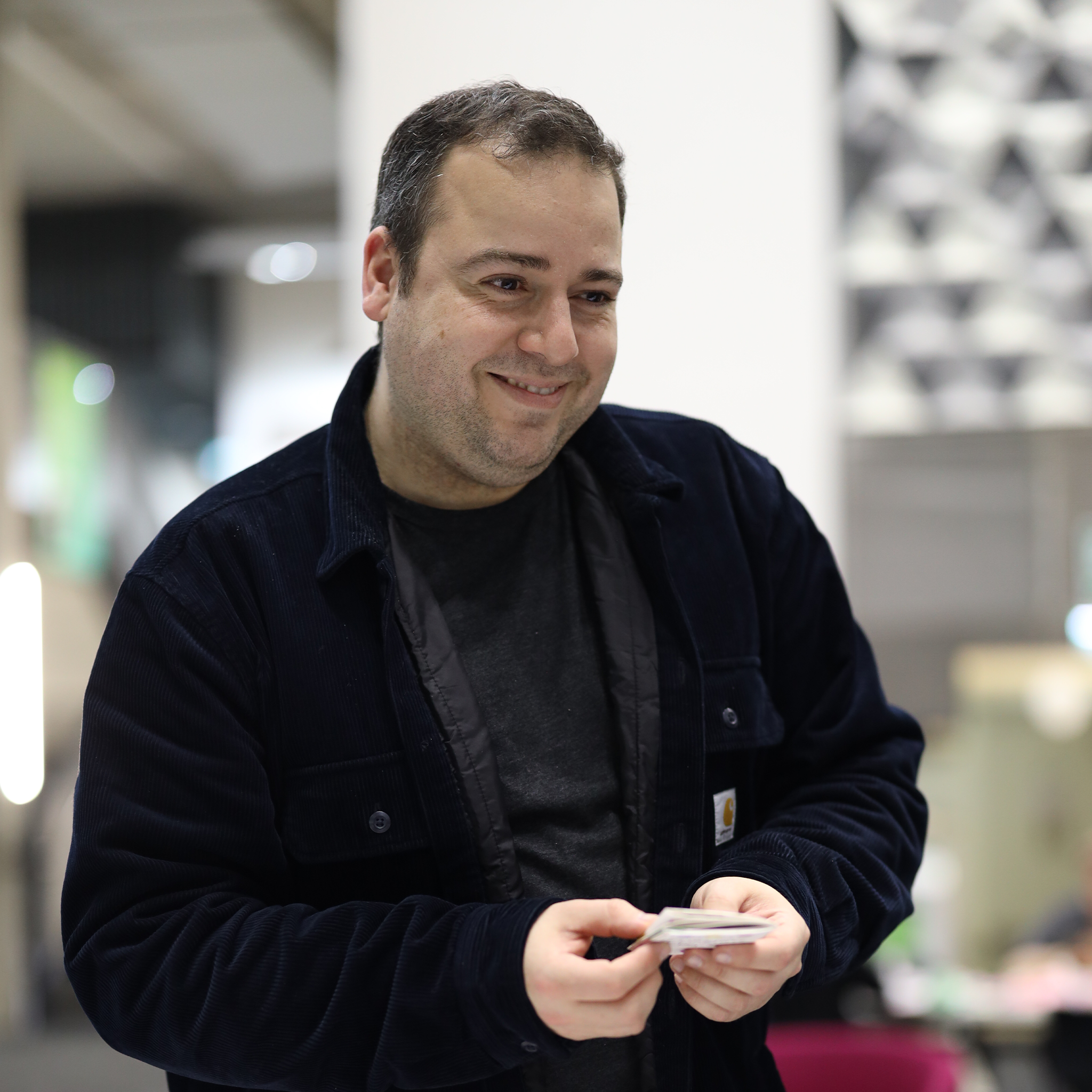 A man with short brown hair, light skin, and wearing a dark jacket over a black shirt, smiling and holding a deck of photos or cards in his hands, standing in an indoor space with a blurred background.