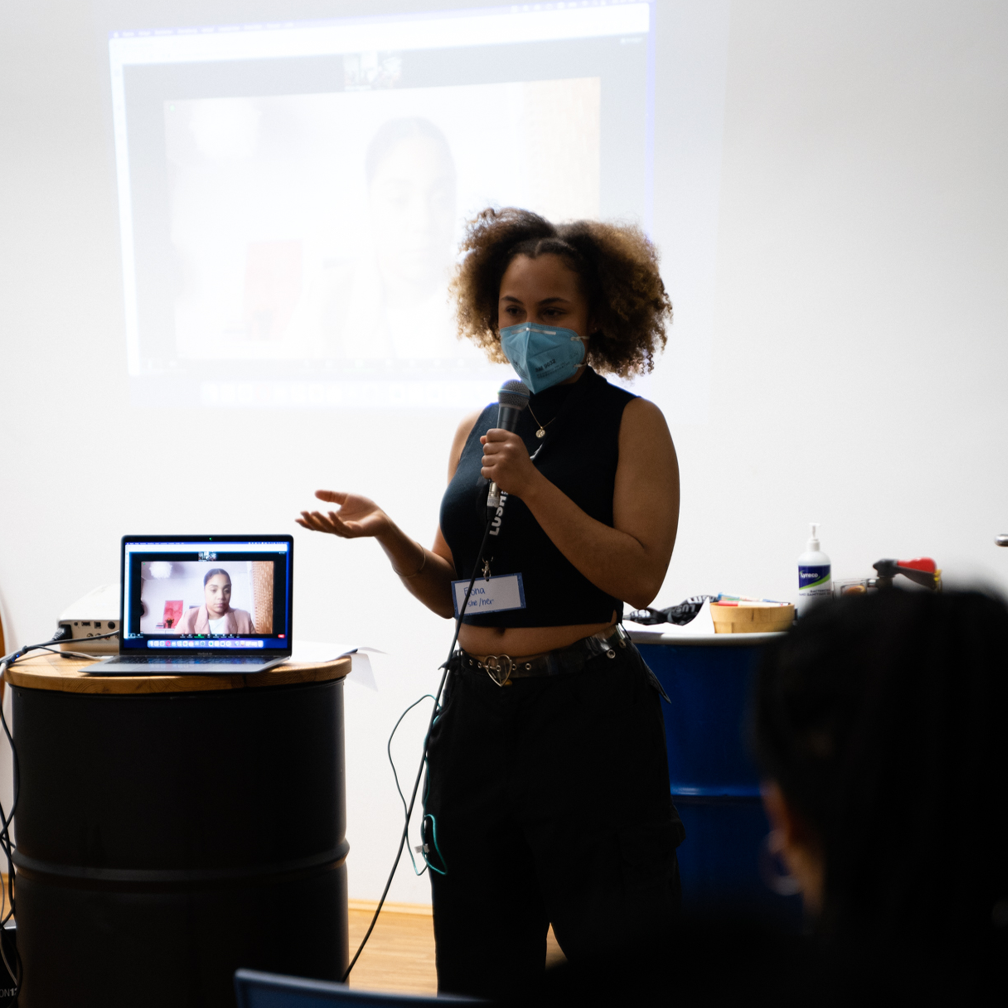 A woman wearing a mask and black sleeveless top giving a presentation in a room with a white wall, with a projector screen behind her showing her image on a computer.