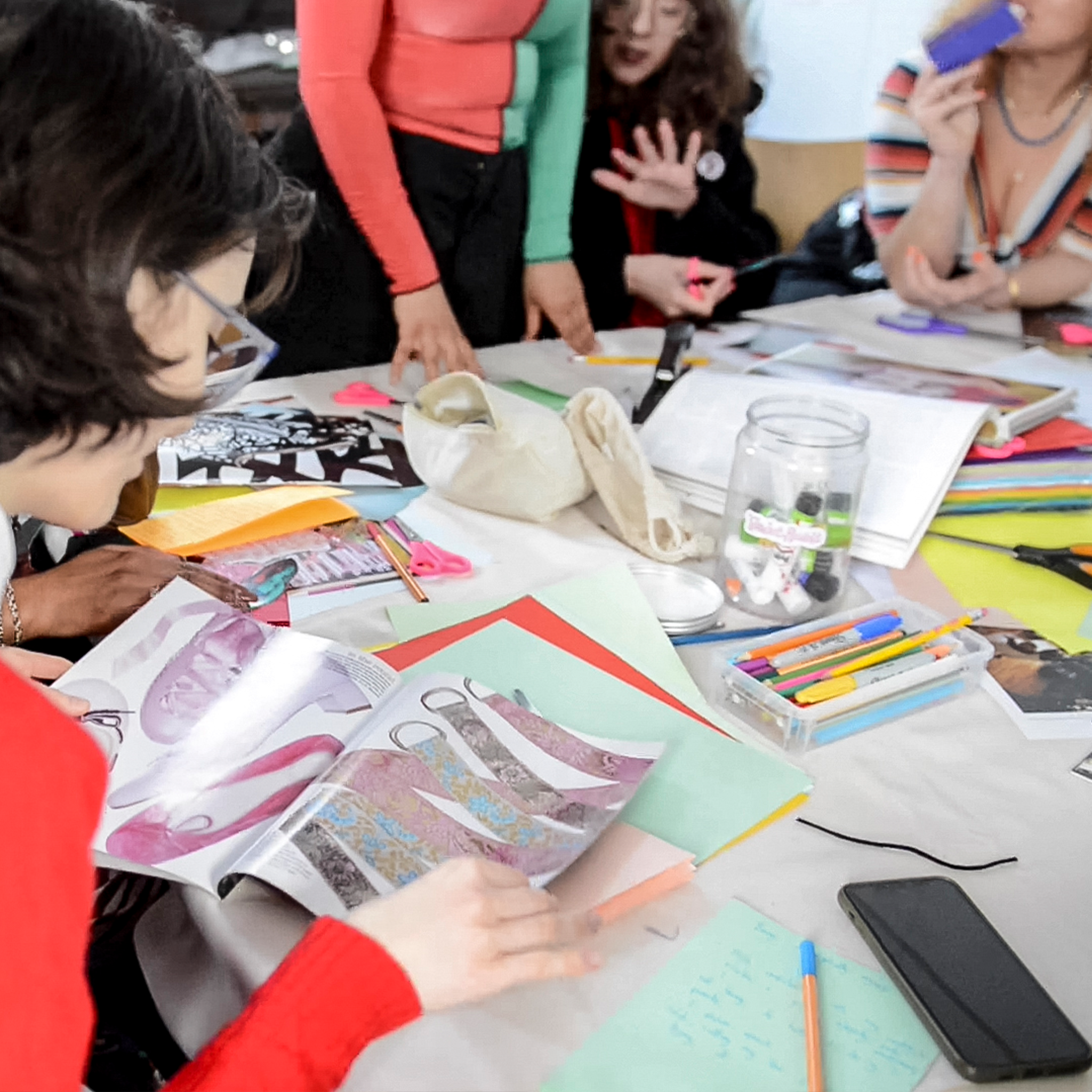 Group of women gathered around a table with magazines, colored pens, scissors, and paper, participating in a creative or crafting activity.