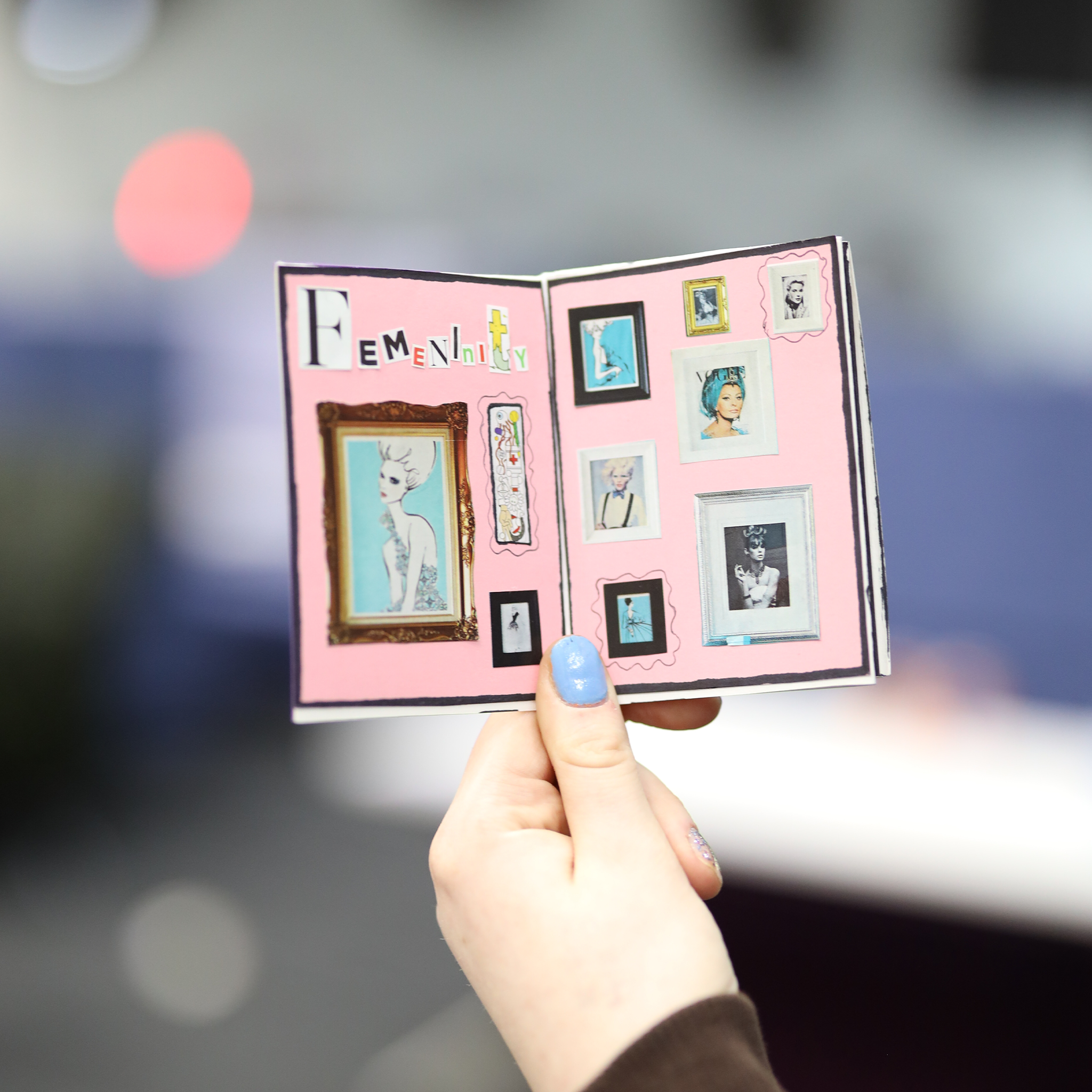 Close-up of a hand holding a pink scrapbook page with collage photographs and artwork related to female empowerment, titled 'Femininty'.