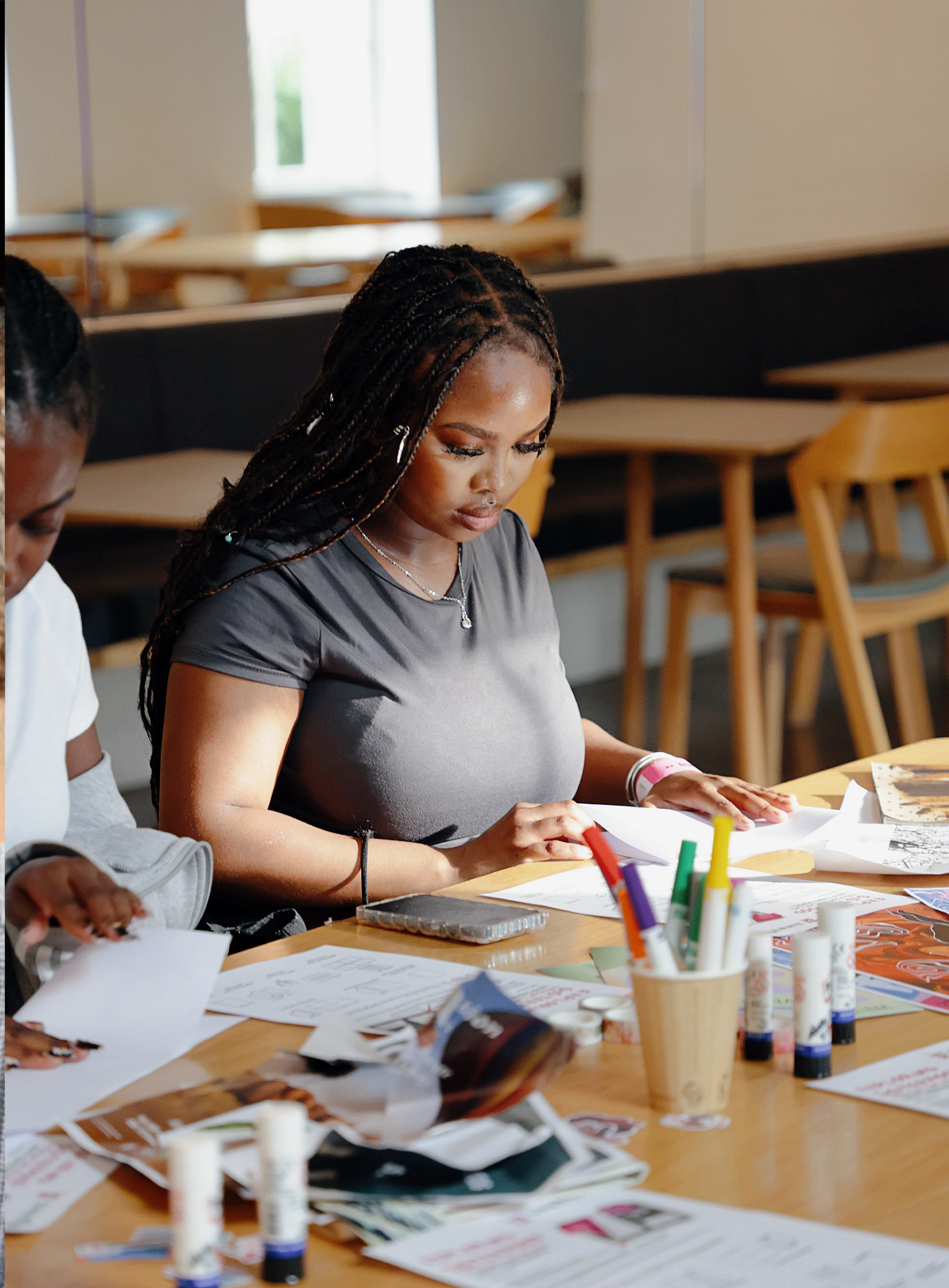 A young woman with long braided hair is sitting at a table looking at papers and surrounded by colorful markers, glue sticks, and flyers in a well-lit room.