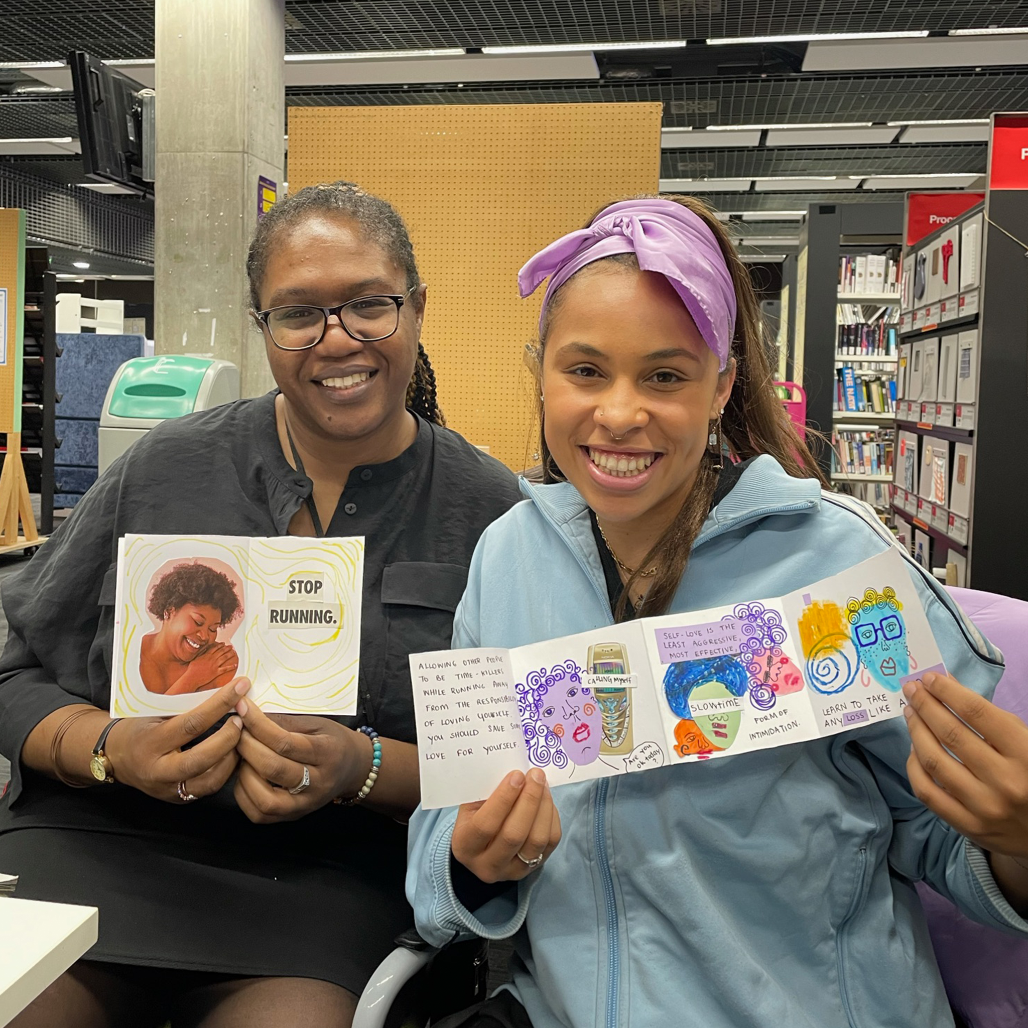 Two women sitting in a bookstore, smiling and holding up colorful illustrated notes about self-love and emotional health.