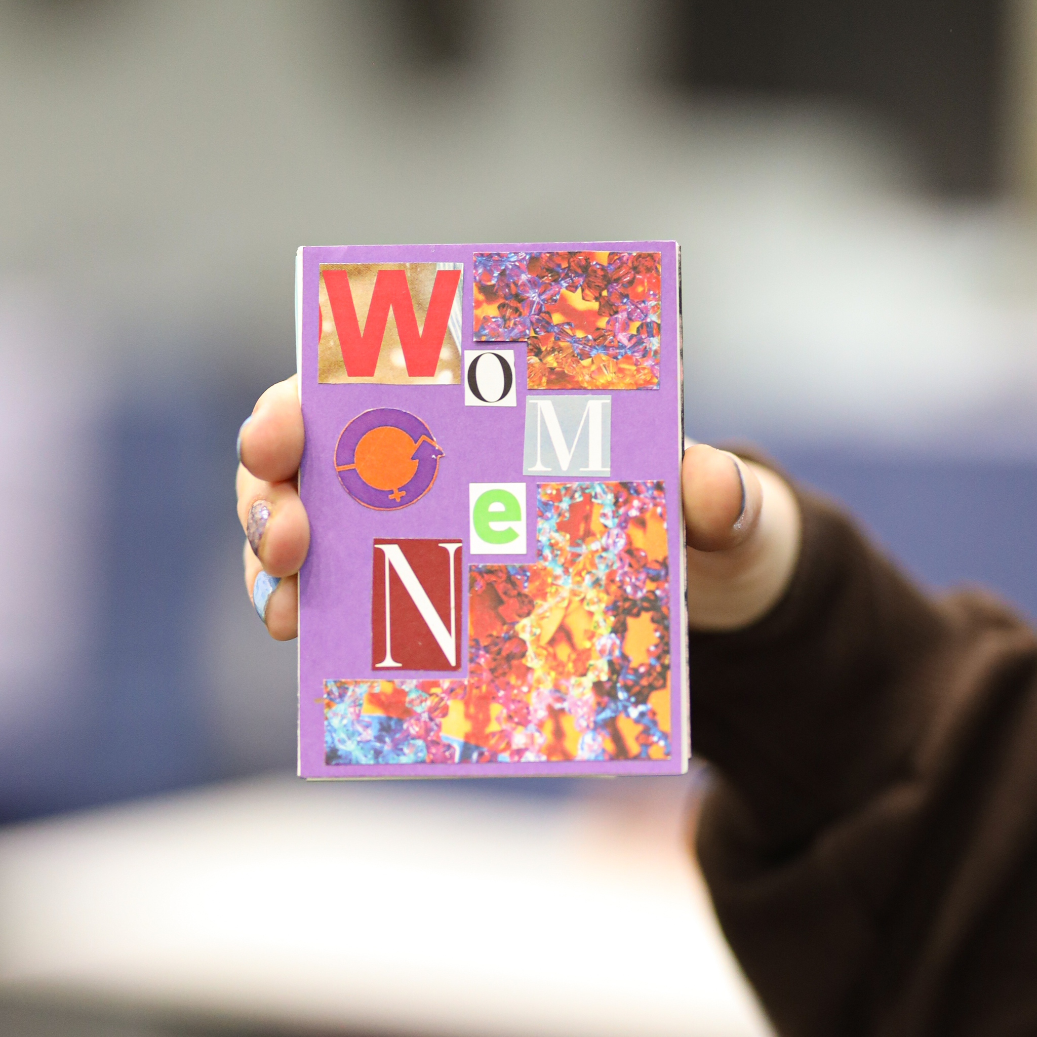 A hand holding a purple card with colorful collage letters spelling 'WOMEN' and 'ONE', with some growth symbols.
