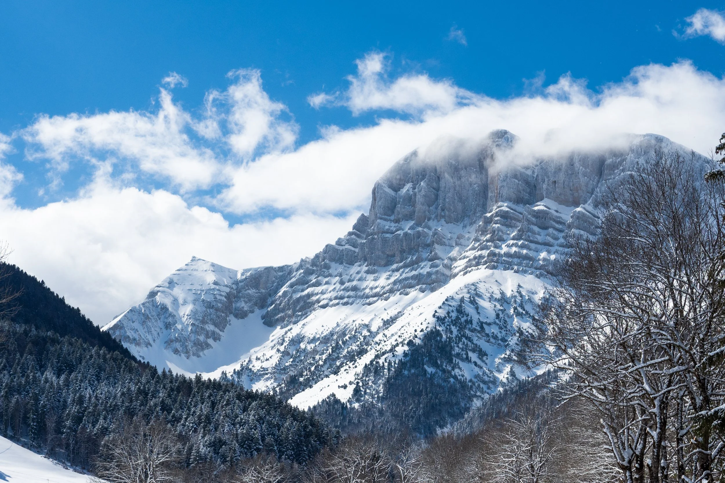 Montagne enneigée avec des sommets élevés et des nuages entourant le sommet, forêt enneigée au premier plan, ciel bleu avec quelques nuages.