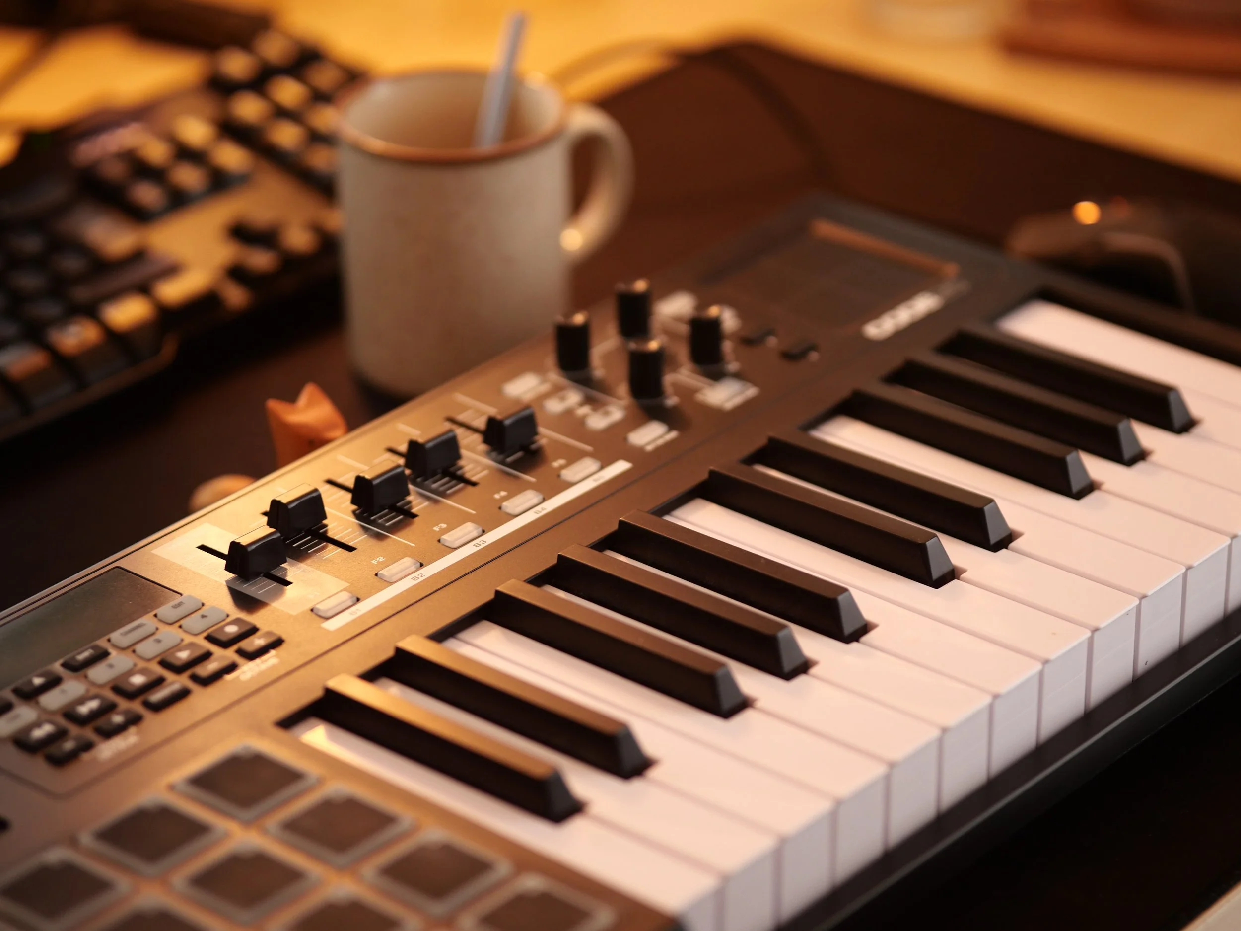 MAO et Musique. Clavier MIDI avec contrôleurs et potards, café dans une tasse, clavier d'ordinateur, et clavier ordinateur en arrière-plan, sur un bureau.