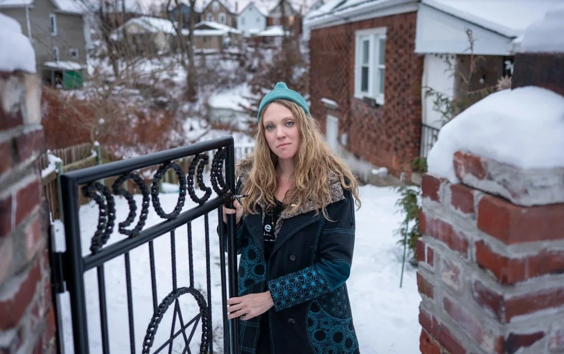 A young woman with long curly blonde hair, wearing a teal beanie and a dark coat, stands next to a black metal gate with a pentagram design, in a snowy residential area with brick houses in the background.