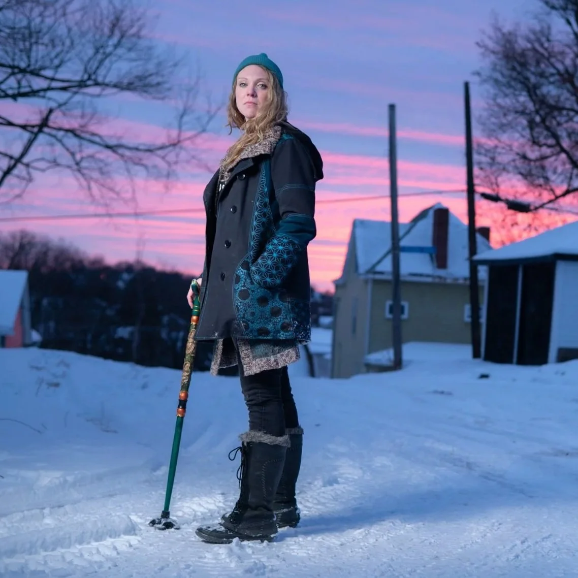 A woman in winter clothing standing on snow with a walking cane, at sunset or sunrise, in a residential area with houses and leafless trees.