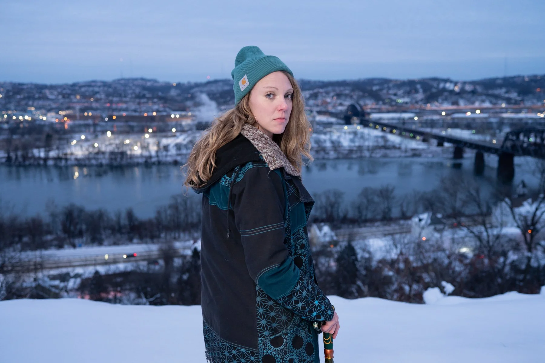 A young woman standing outdoors in winter, wearing a teal beanie and a black jacket, with a snowy landscape and a river with a bridge in the background.