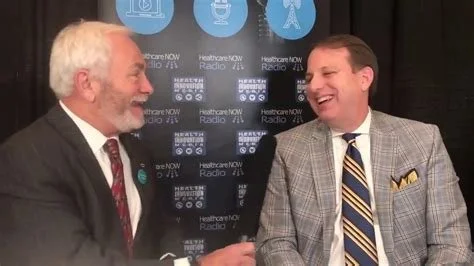 Two men are talking and smiling during a radio interview in front of a backdrop with HealthcareNOW Radio logos. One man has white hair and a beard, wearing a dark suit; the other has short brown hair, wearing a gray checkered suit and striped tie.