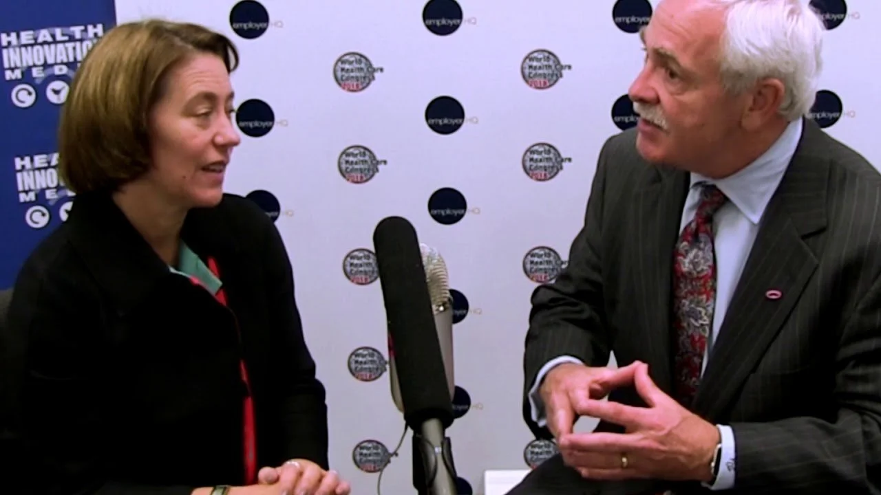 Two individuals engaged in a conversation during an interview, with a backdrop containing logos related to health and innovation.