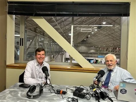 Two men sitting at a table with microphones in a radio studio or recording booth, with a large window behind them showing an industrial or warehouse-like interior.