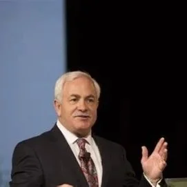 An older man with white hair, wearing a dark suit and a light-colored shirt, standing in front of a blue and black background, speaking or presenting.