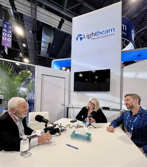 Three people sitting at a table with microphones, a woman in the middle and two men, at a Lightbeam Health Solutions booth in an exhibition hall or conference center.