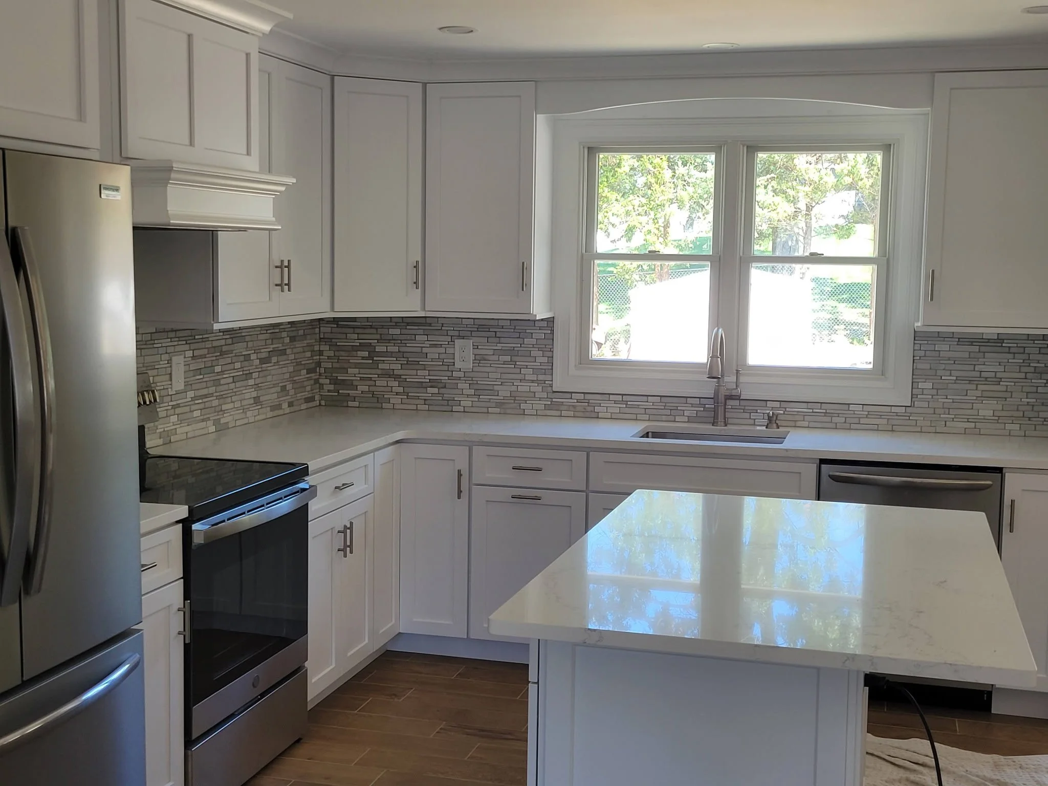 Modern kitchen with white cabinets, stainless steel appliances, and a large window over the sink, with a light-colored countertop and wood flooring.