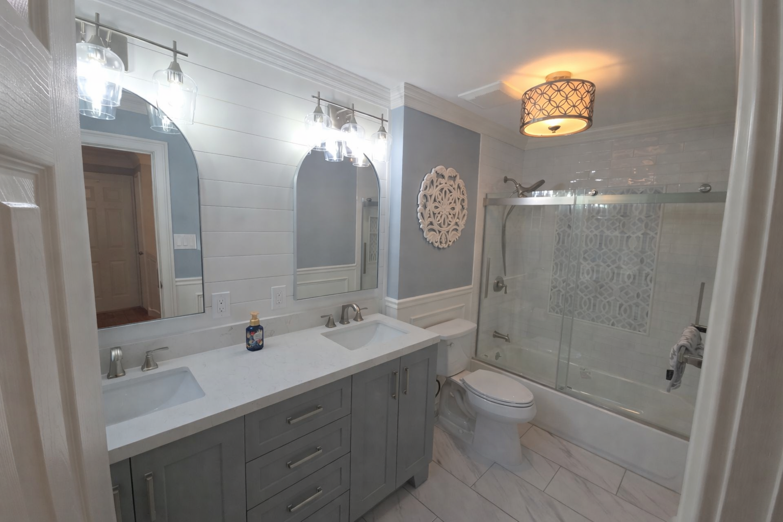 Modern bathroom featuring a double sink vanity with a white countertop, two mirrors, and wall-mounted lights. There is a toilet next to a glass-enclosed shower with decorative wall tile.