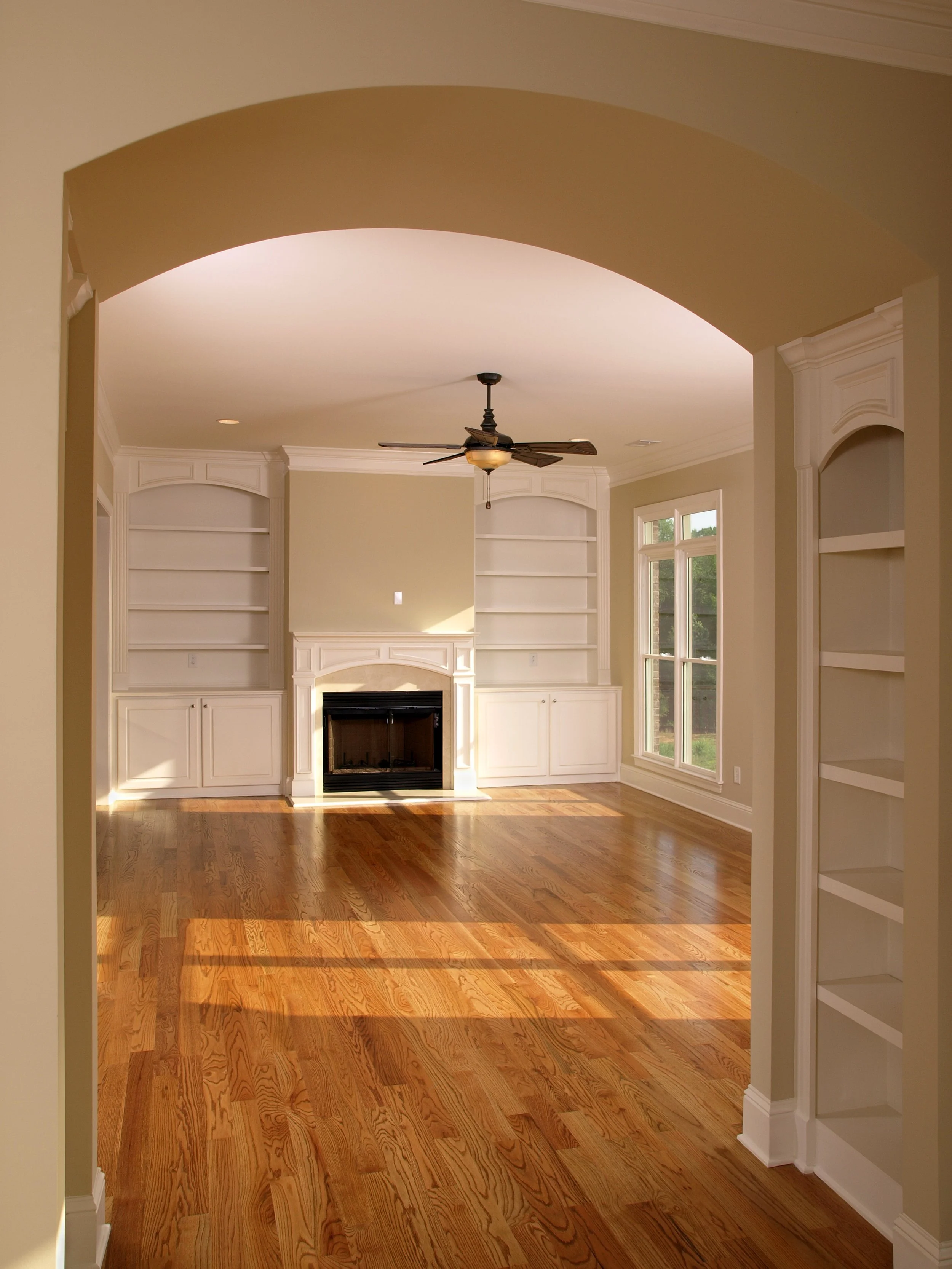 Empty living room with wooden floors, built-in white bookshelves, a fireplace, large window, and ceiling fan.