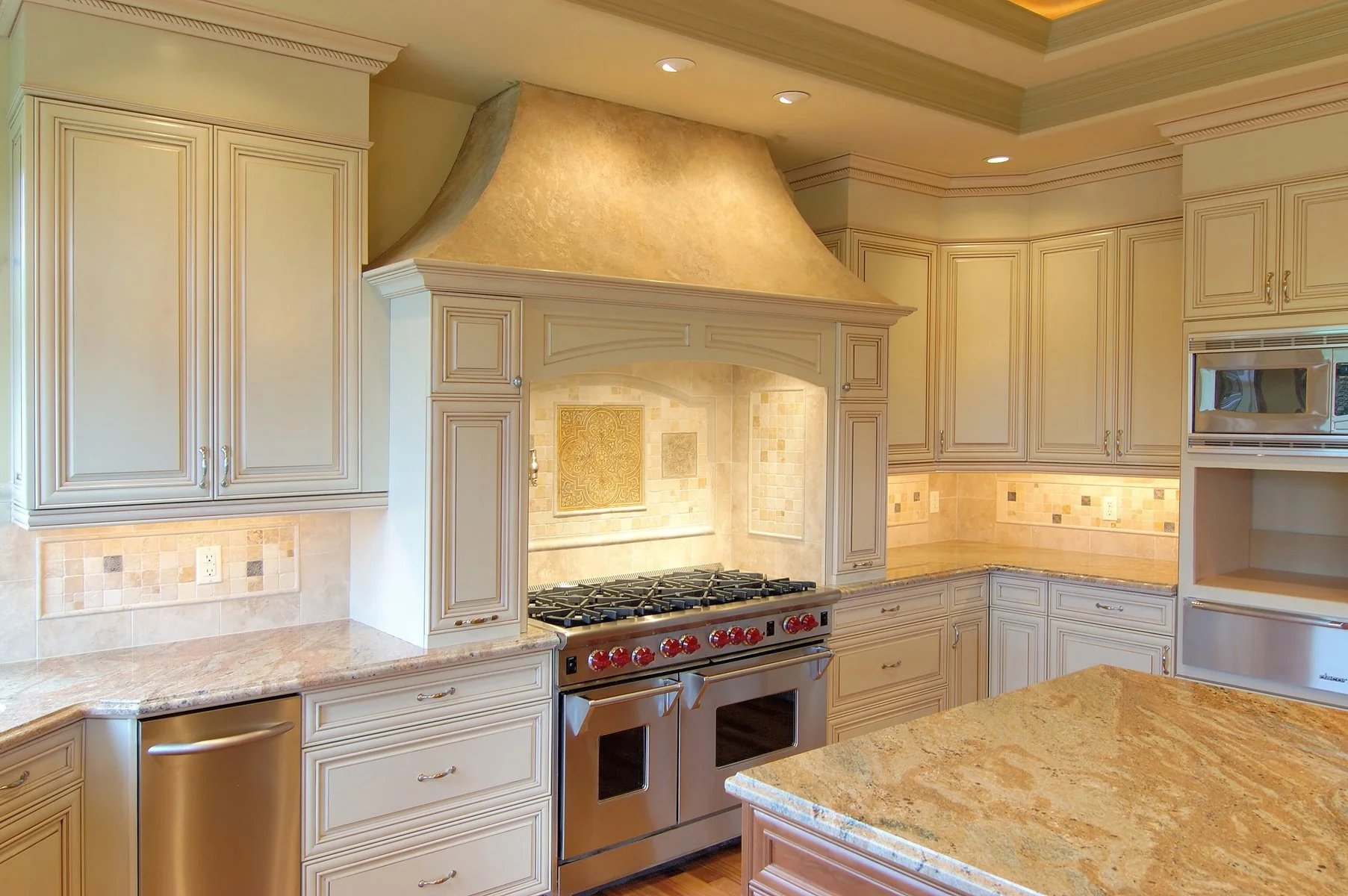 Kitchen with cream-colored cabinets, granite countertops, a large stainless steel stove with red knobs, decorative backsplash tiles, and an ornate range hood.