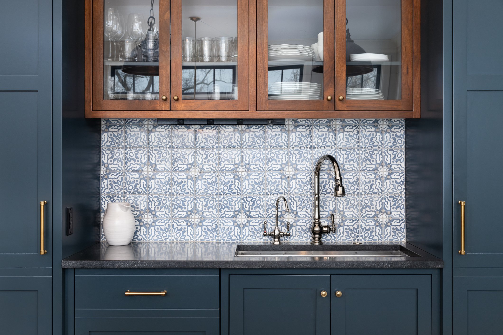 A kitchen sink area with blue cabinetry, a gray countertop, and a patterned tile backsplash. There are wooden cabinets with glass doors above, displaying glassware and dishes. The sink has two faucets, and a white vase is placed on the left side of the counter.
