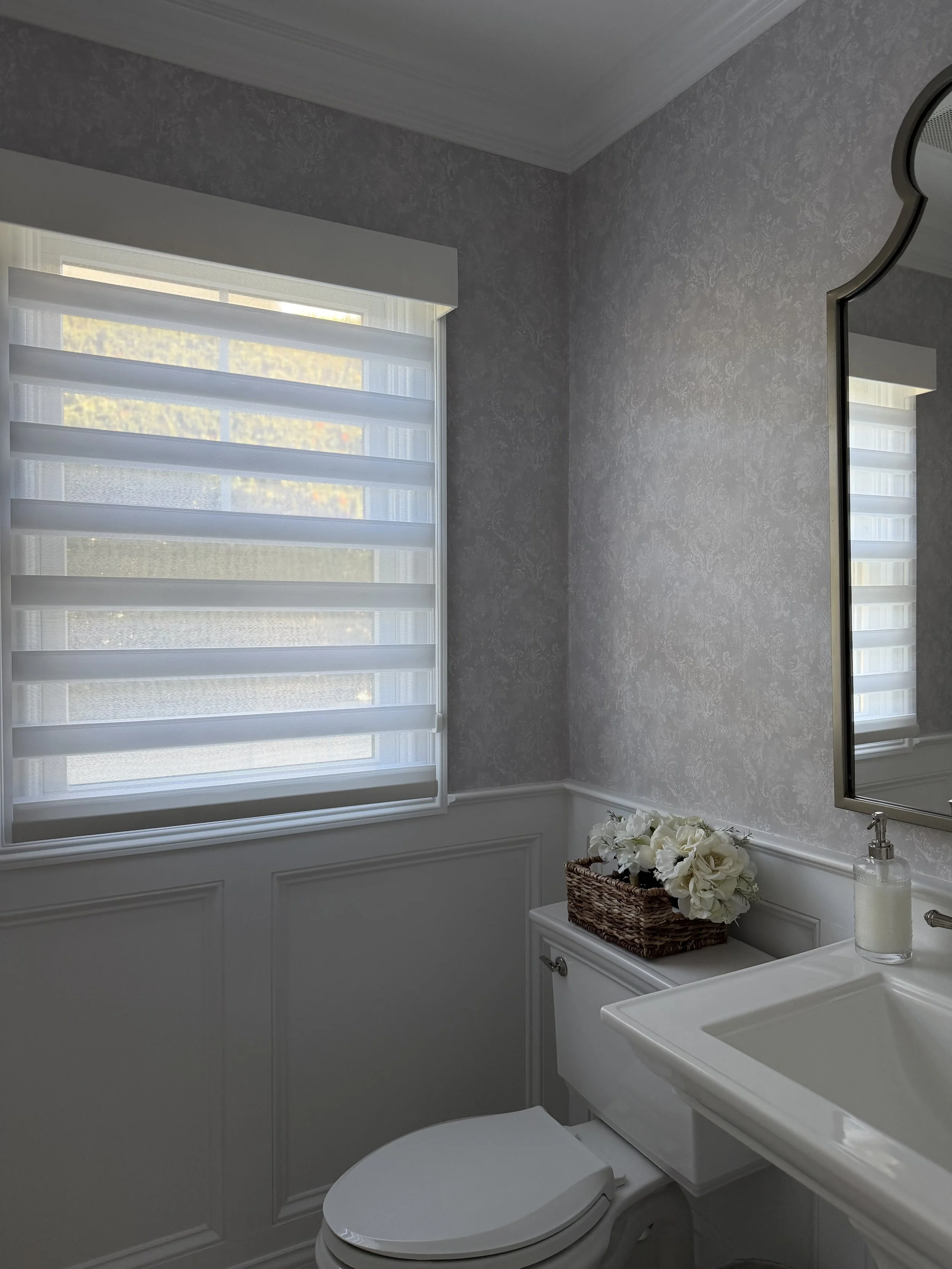 A bathroom with a window covered by white horizontal blinds, a wainscoting wall, a sink with a soap dispenser, and a decorative basket with white flowers on the toilet tank.