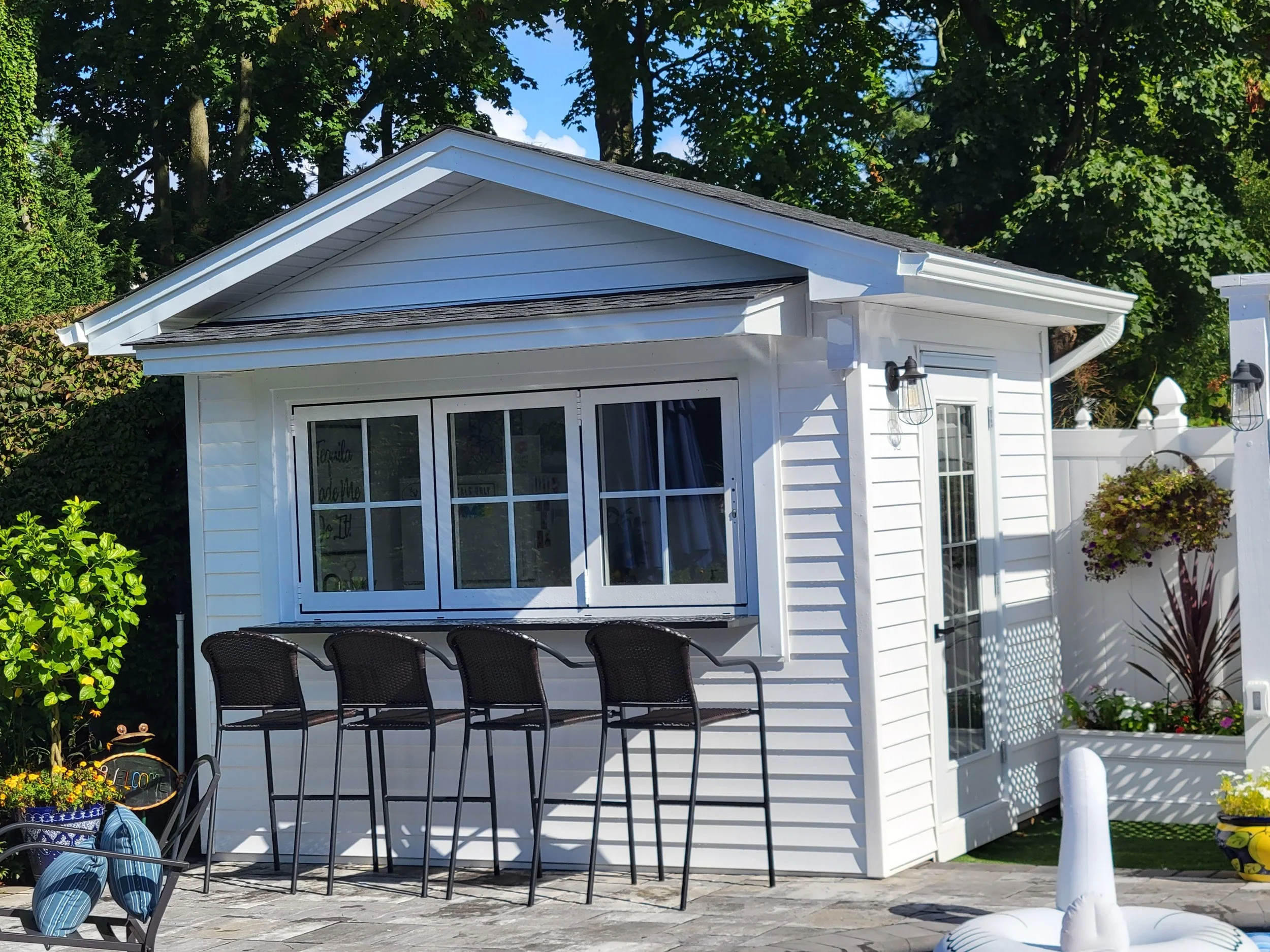 White backyard poolhouse with four black barstools outside, surrounded by green trees and decorated with plants and hanging flower baskets, on a sunny day.