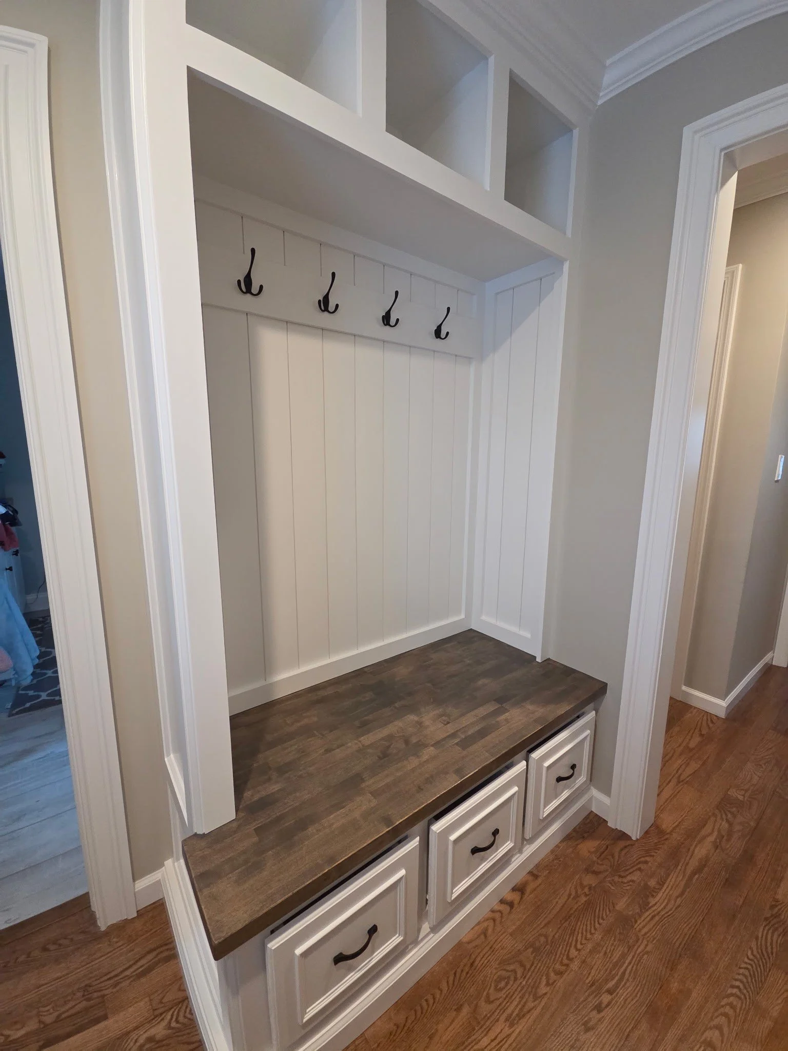 A built-in entryway storage bench with white drawers, a dark wooden top, and hooks for hanging items, located in a home interior with hardwood floors and light-colored walls.