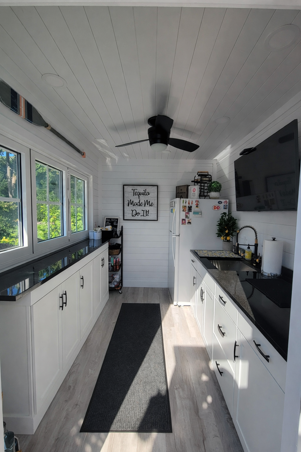 Bright white kitchen with black countertops, large window, and a black ceiling fan. There's a white fridge with magnets, a black sink with a potted plant, a wall-mounted TV, and a framed sign that says 'Tequila Made Me Do It!'. Light wood flooring and a black runner rug on the floor.