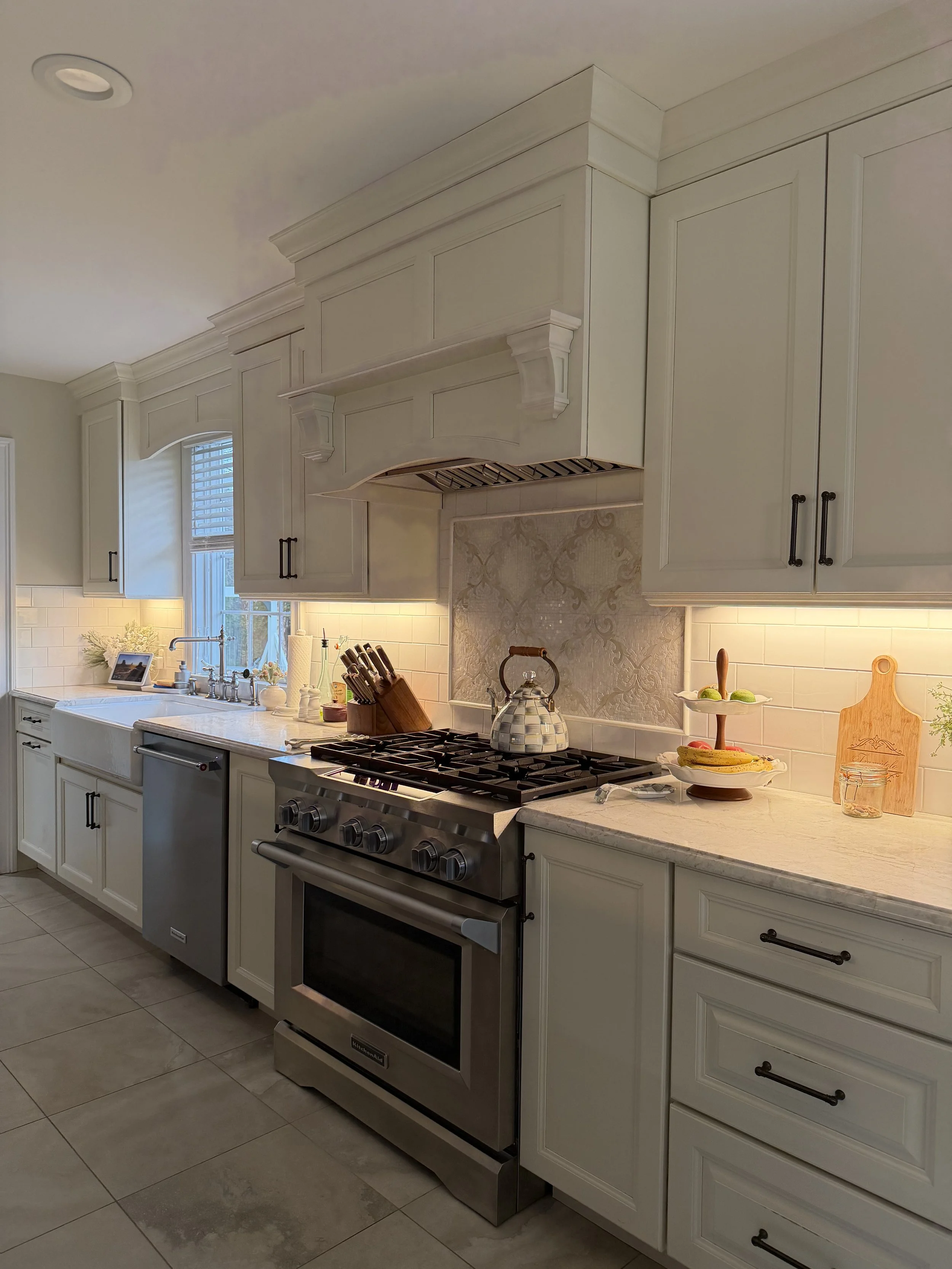 Modern kitchen with white cabinets, stainless steel stove, teapot on stove, countertop with cutting board, jars, and tiered tray with gourds, and a window near the sink.