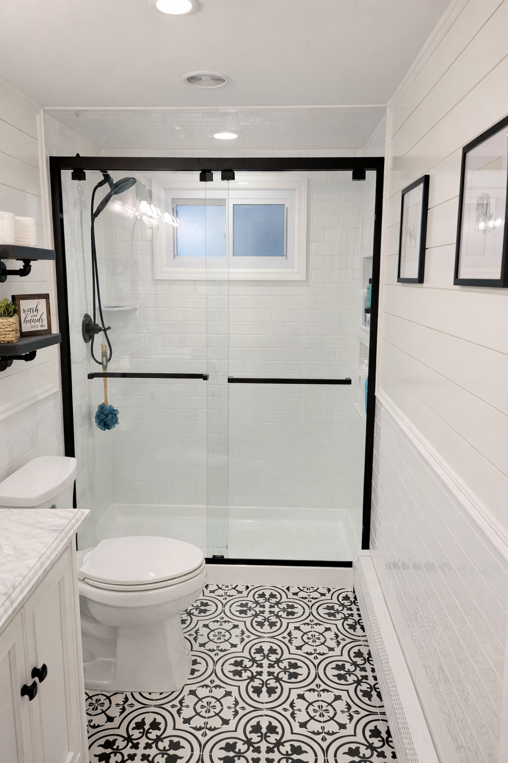 Bathroom with a glass shower enclosure, black metal frame, white tile walls, patterned black and white floor tiles, toilet, and wall-mounted shelves with decorative items.