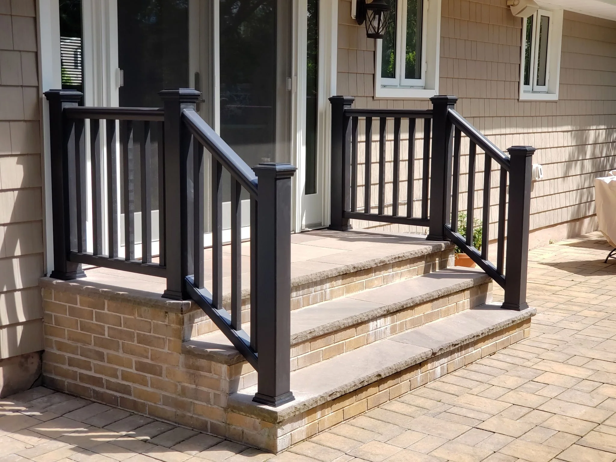 A small porch with brick steps and black wooden railings attached to a house with beige siding and white window frames.