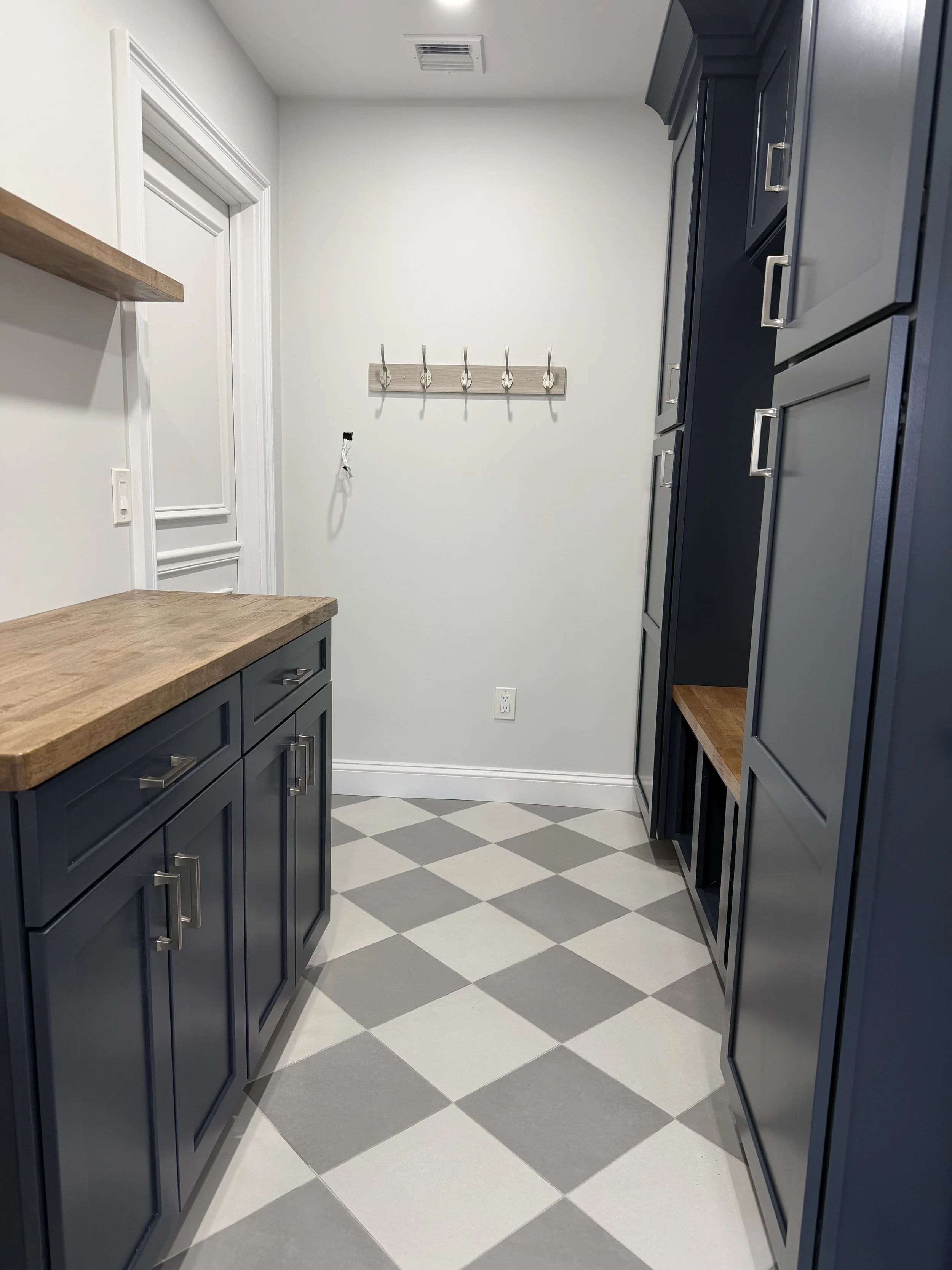 A small laundry room with blue cabinets, wooden countertops, a wooden wall hook strip, white walls, checkered gray and white tile floor, and a white door.