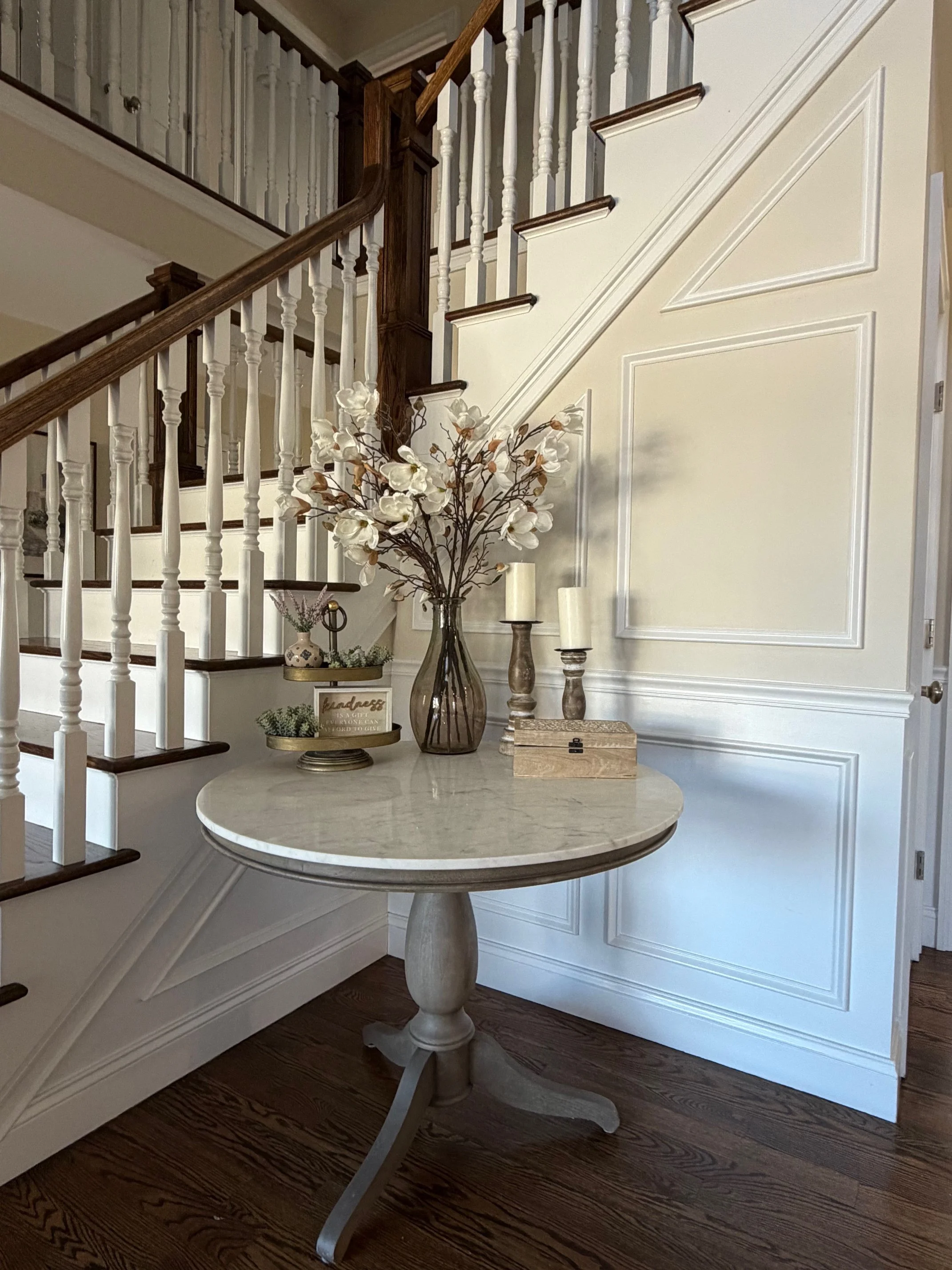 Decorative table with flower arrangement, candles, and small decorative items near staircase in a home interior.