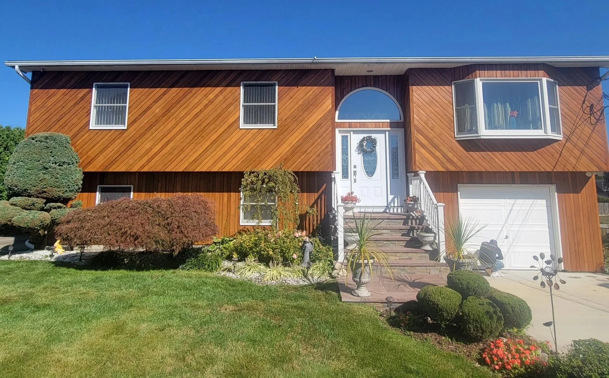 A two-story house with a wood-paneled exterior, a white front door decorated with a wreath, a small set of steps, and a landscaped front yard with shrubs, potted plants, and a green lawn.
