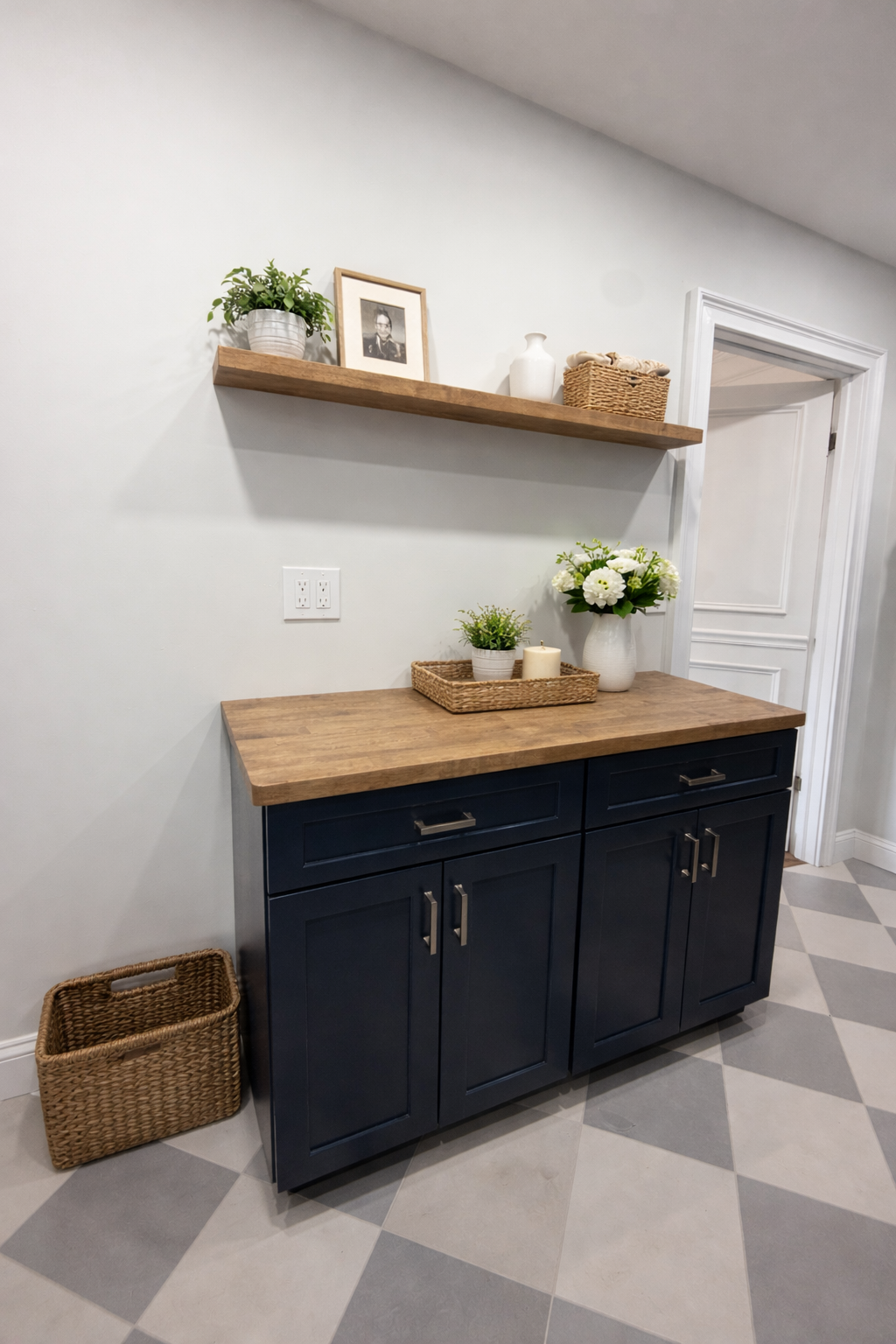 A kitchen cabinet with a wooden countertop, decorated with a basket, candles, potted plants, and a white vase with flowers. A wooden shelf above holds a potted plant, framed photo, and decorative objects. The floor has a checkered pattern of light an