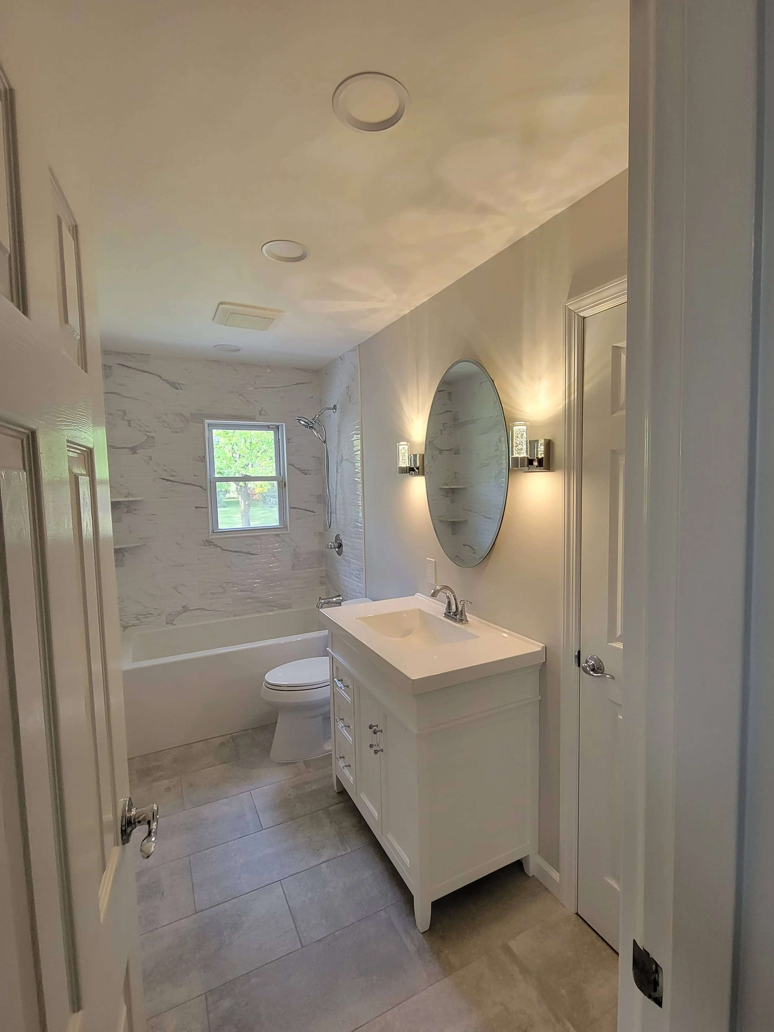 A modern bathroom with a bathtub and shower area, a toilet, a white vanity with a sink, a large oval mirror, and wall-mounted lights. The bathroom has light-colored walls, tile flooring, and a small window in the shower area.