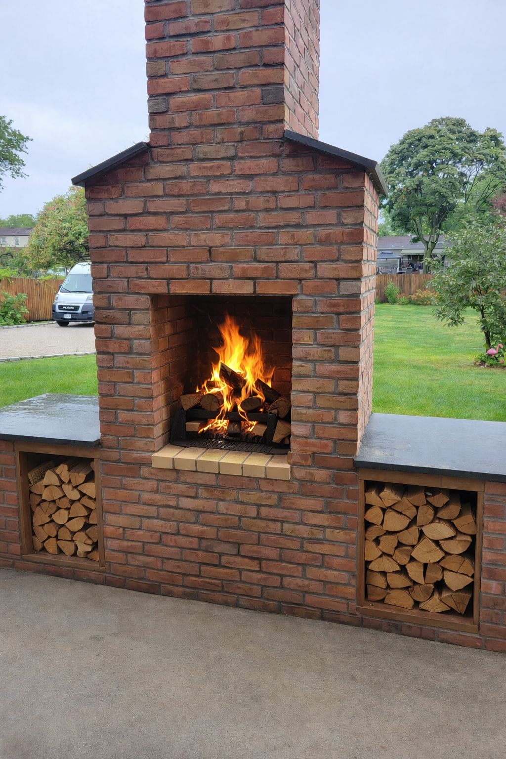 Outdoor brick fireplace with a fire burning, flanked by wood storage compartments filled with chopped firewood, in a backyard with a grassy lawn and trees.