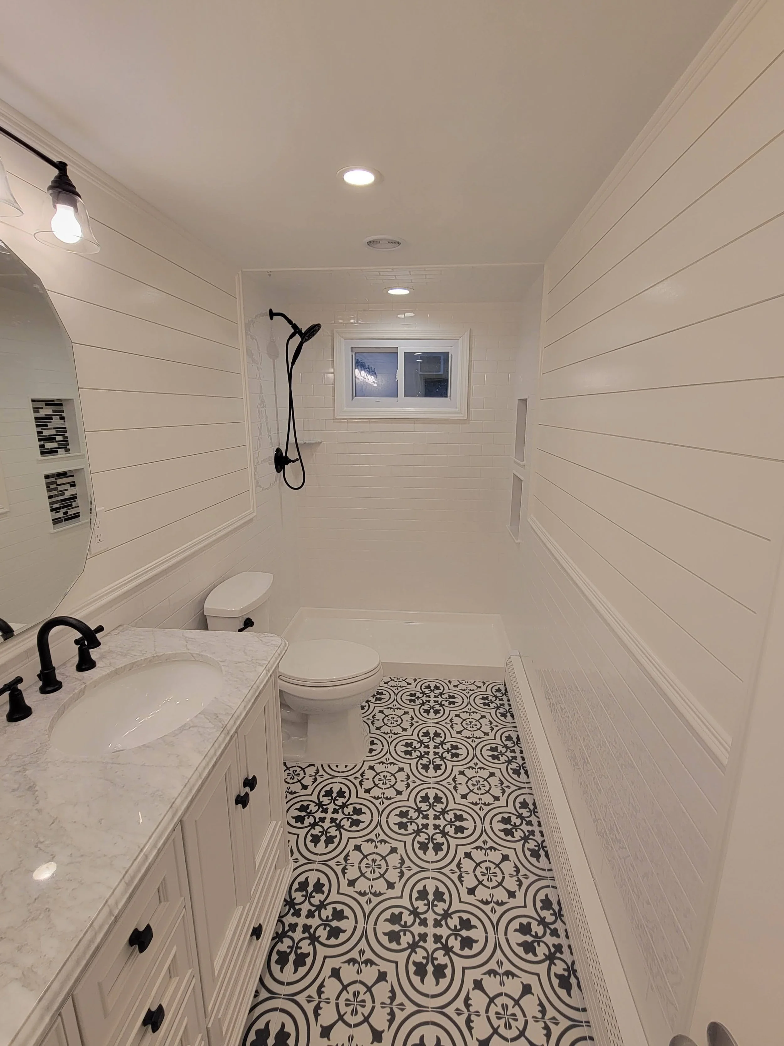 Bathroom with black and white patterned tile flooring, white shiplap walls, a small window, a shower with a black handheld shower head, white vanity with a marble countertop, a round mirror, and ceiling lights.