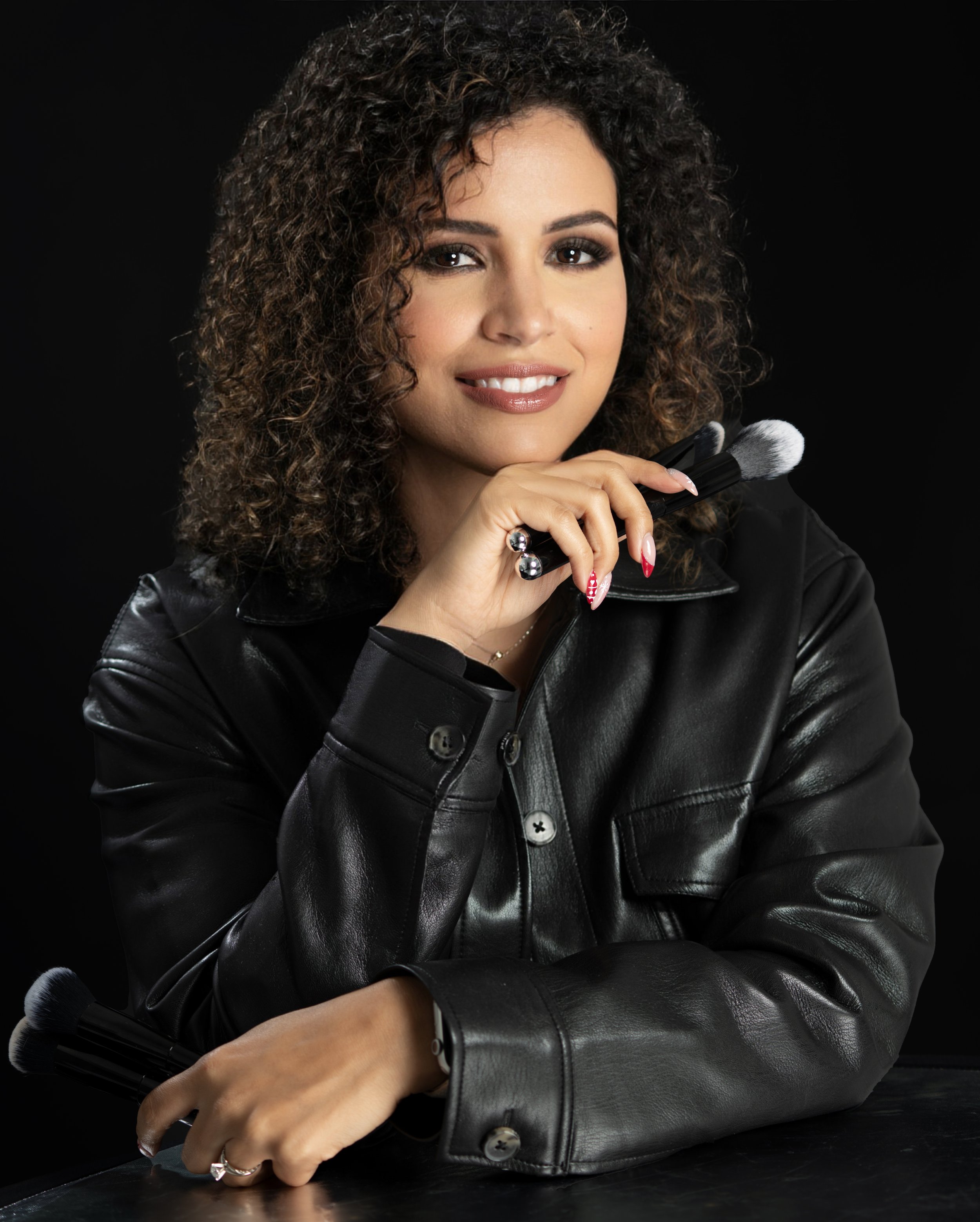 A woman with curly brown hair, wearing a black leather jacket, holding makeup brushes, smiling, against a black background.