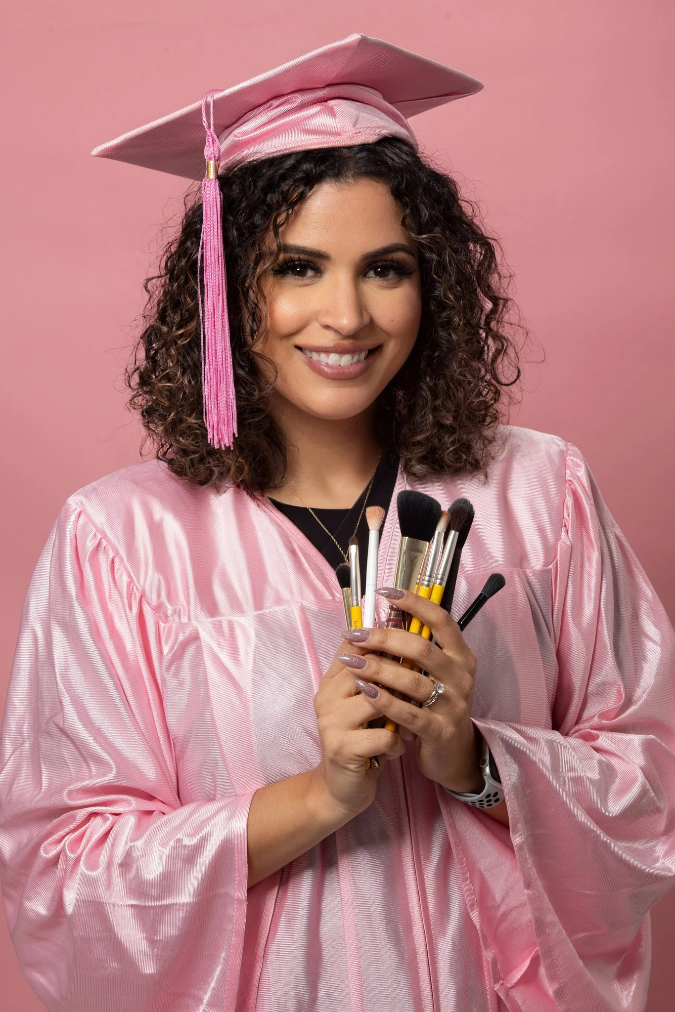 A woman in a pink graduation gown and cap holds a bunch of makeup brushes.