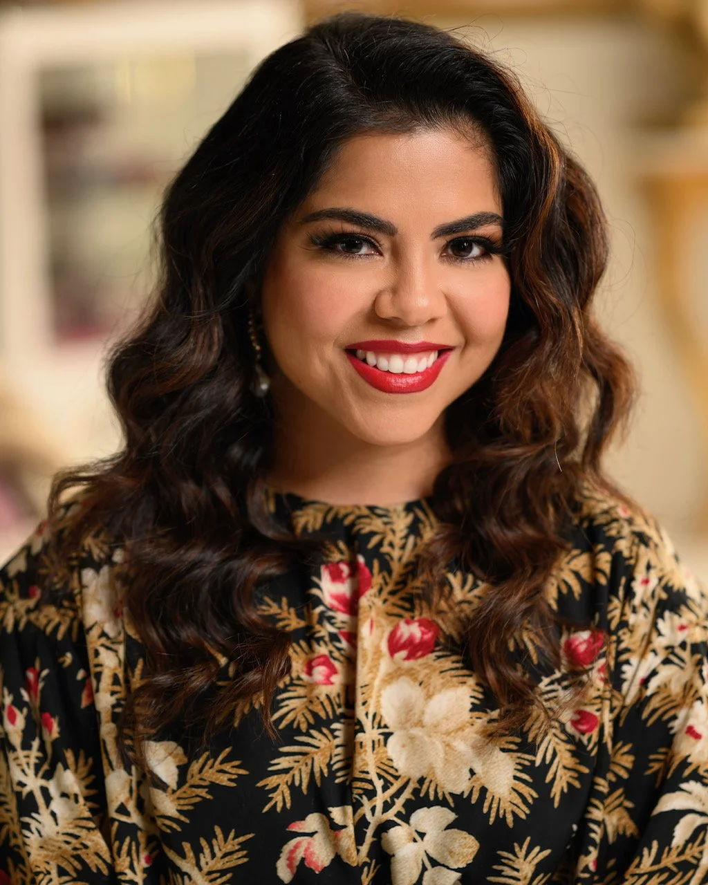 A woman with dark, wavy hair and bright red lipstick, wearing a floral patterned top, smiling at the camera.
