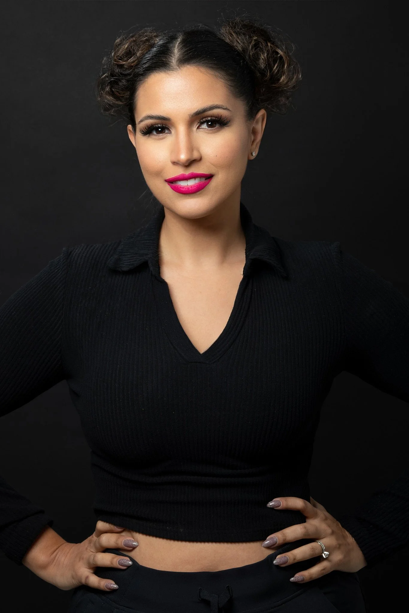 A woman with dark hair styled in two buns, wearing a black V-neck top, black pants, and pink lipstick, standing against a black background with her hands on her hips.