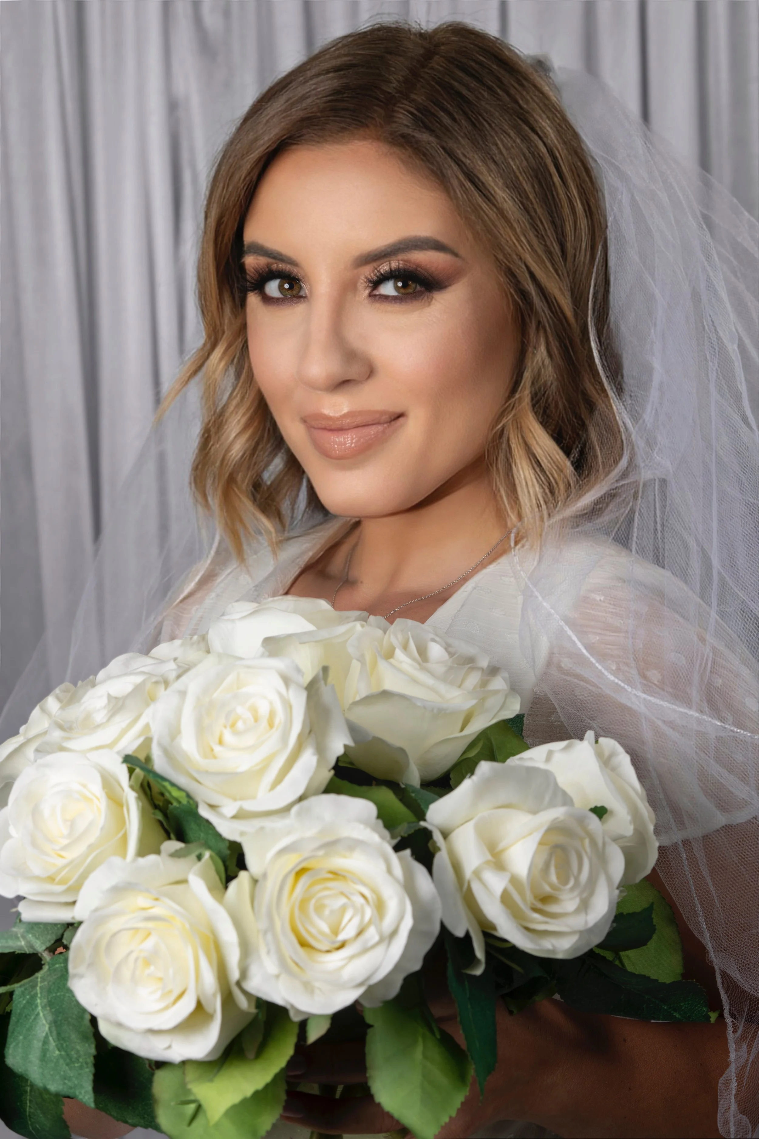 A woman with shoulder-length brown hair, makeup, and a veil, holding a bouquet of white roses, smiling at the camera, with a gray curtain background.
