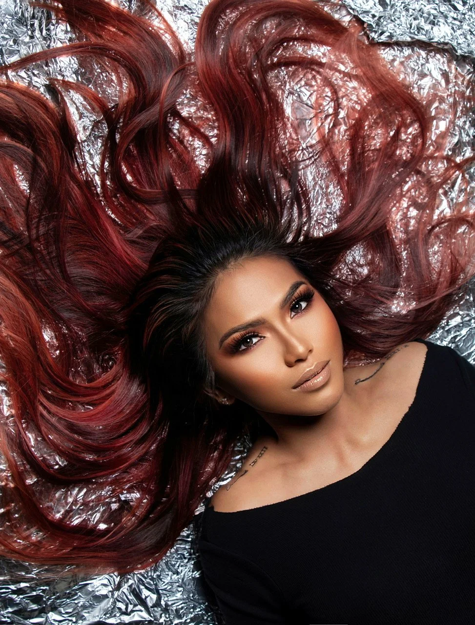 A woman with long, wavy, red and black hair lies on a silver reflective surface, looking at the camera with a neutral expression.