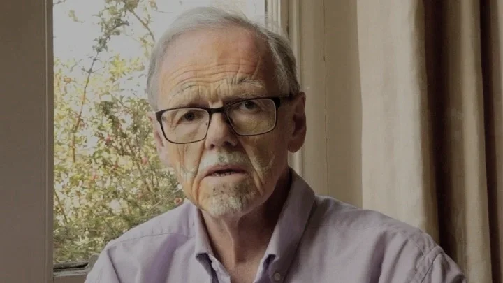 An elderly man with glasses and a light purple shirt looks confused or skeptical, sitting indoors near a window with trees in the background.