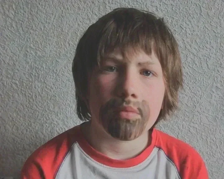 A young man with medium-length brown hair, a painted beard, and mustache, wearing a red and gray shirt, standing against a textured wall.